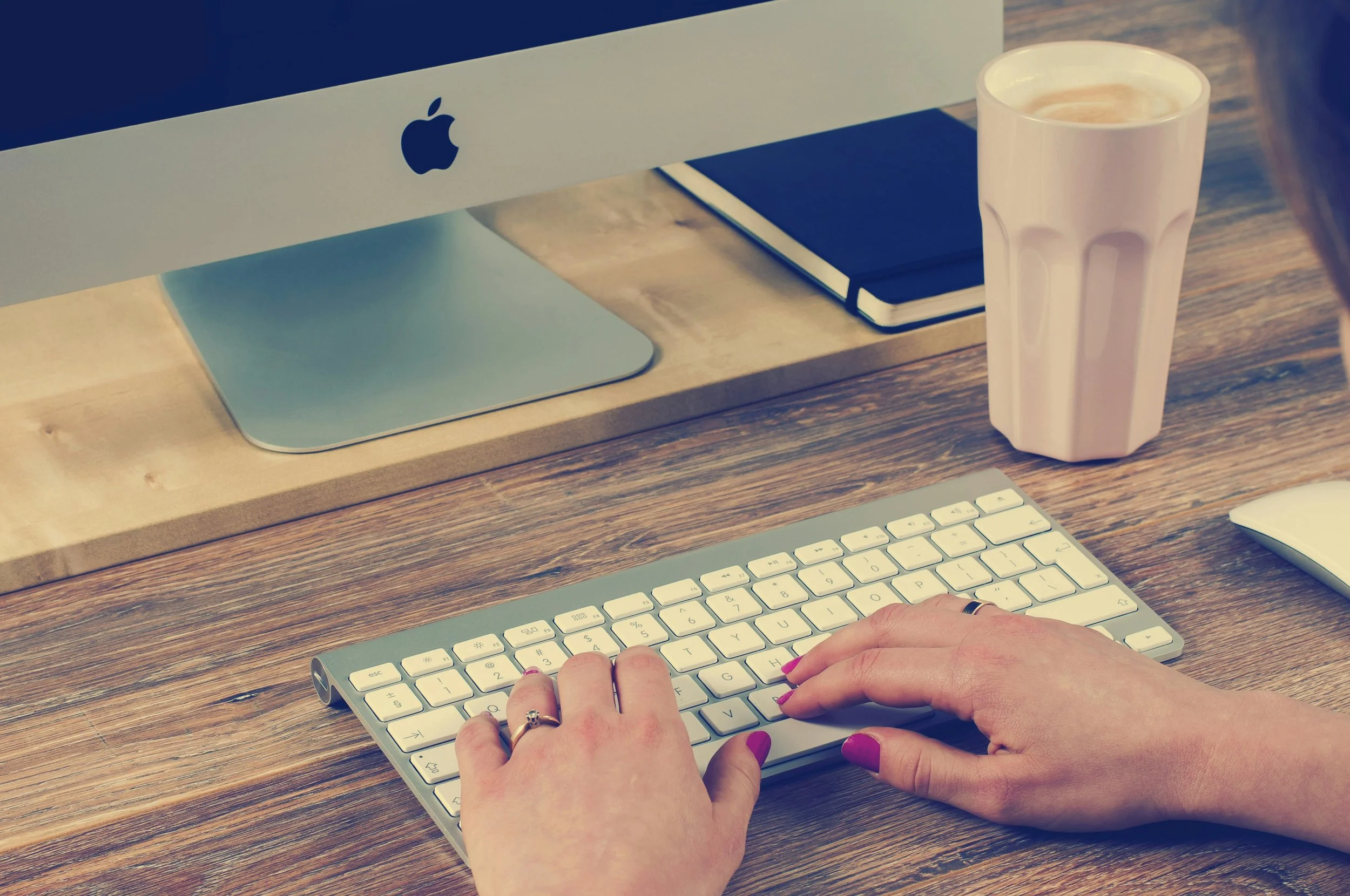 Person typing on a keyboard at a desk with an Apple computer monitor, a notebook, and a pink cup.