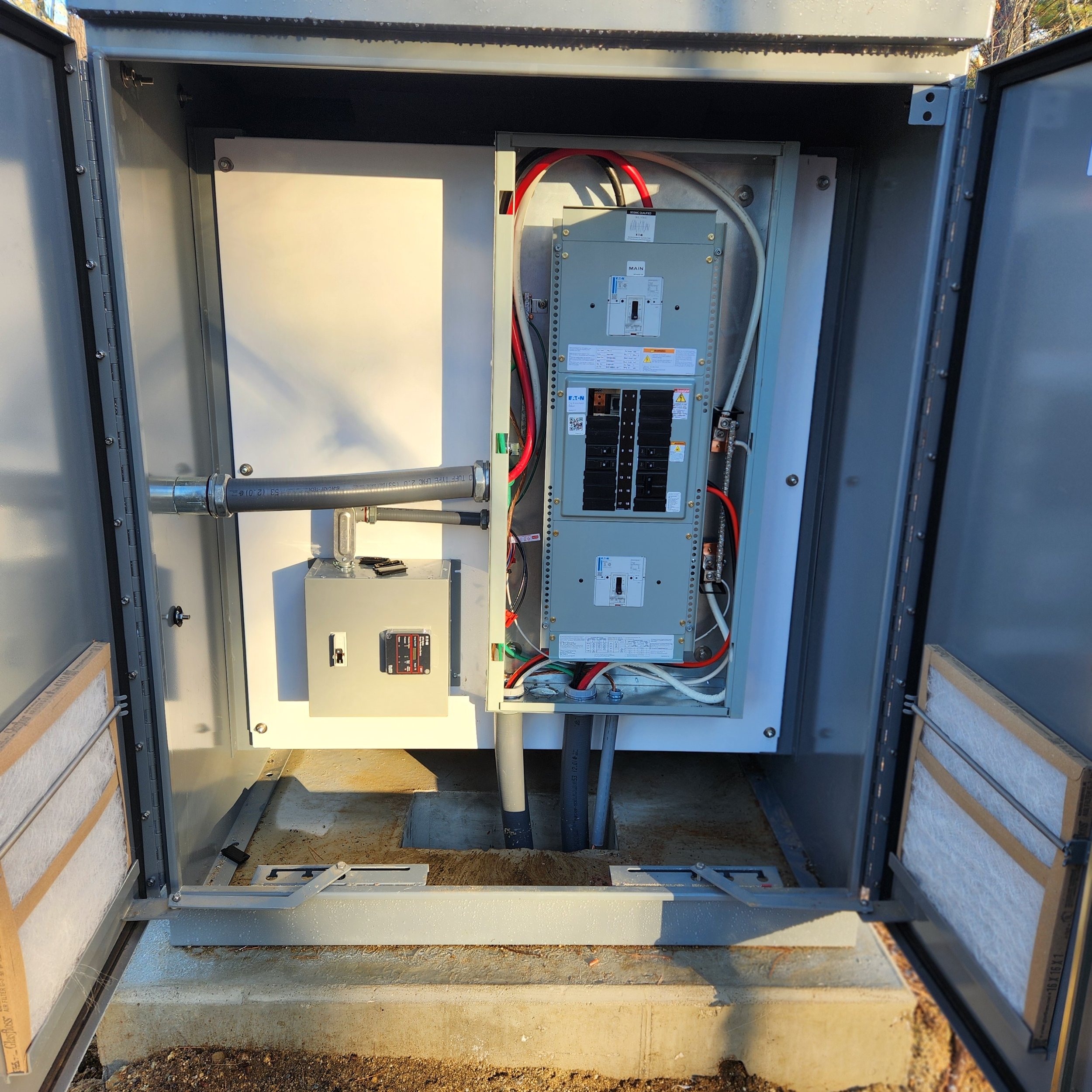 Electrical utility box with circuit breakers and wiring inside an open metal enclosure on a concrete pad.