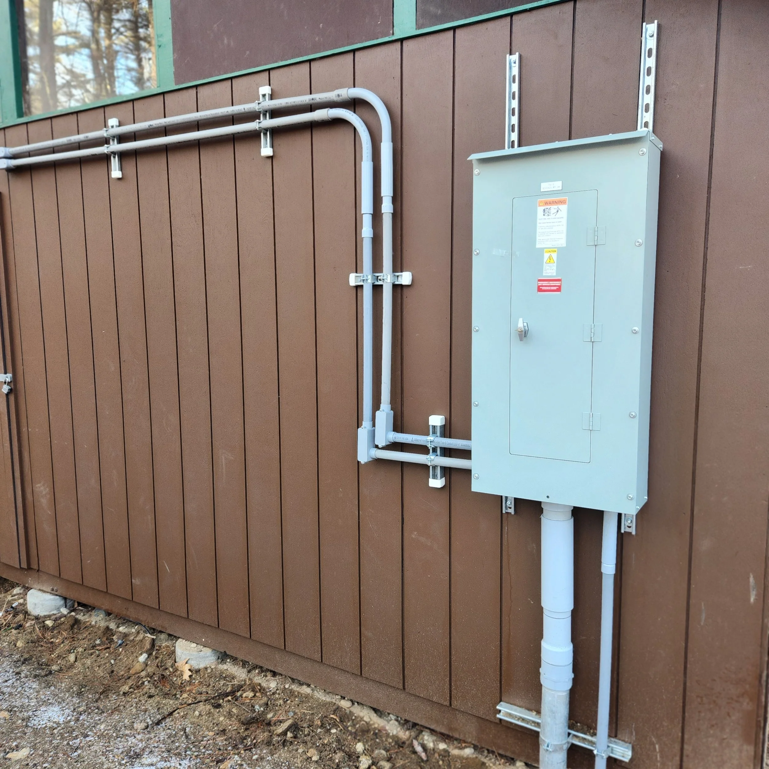 Electrical panel with gray metal door mounted on a brown wooden wall, with electrical conduit pipes connected to it.