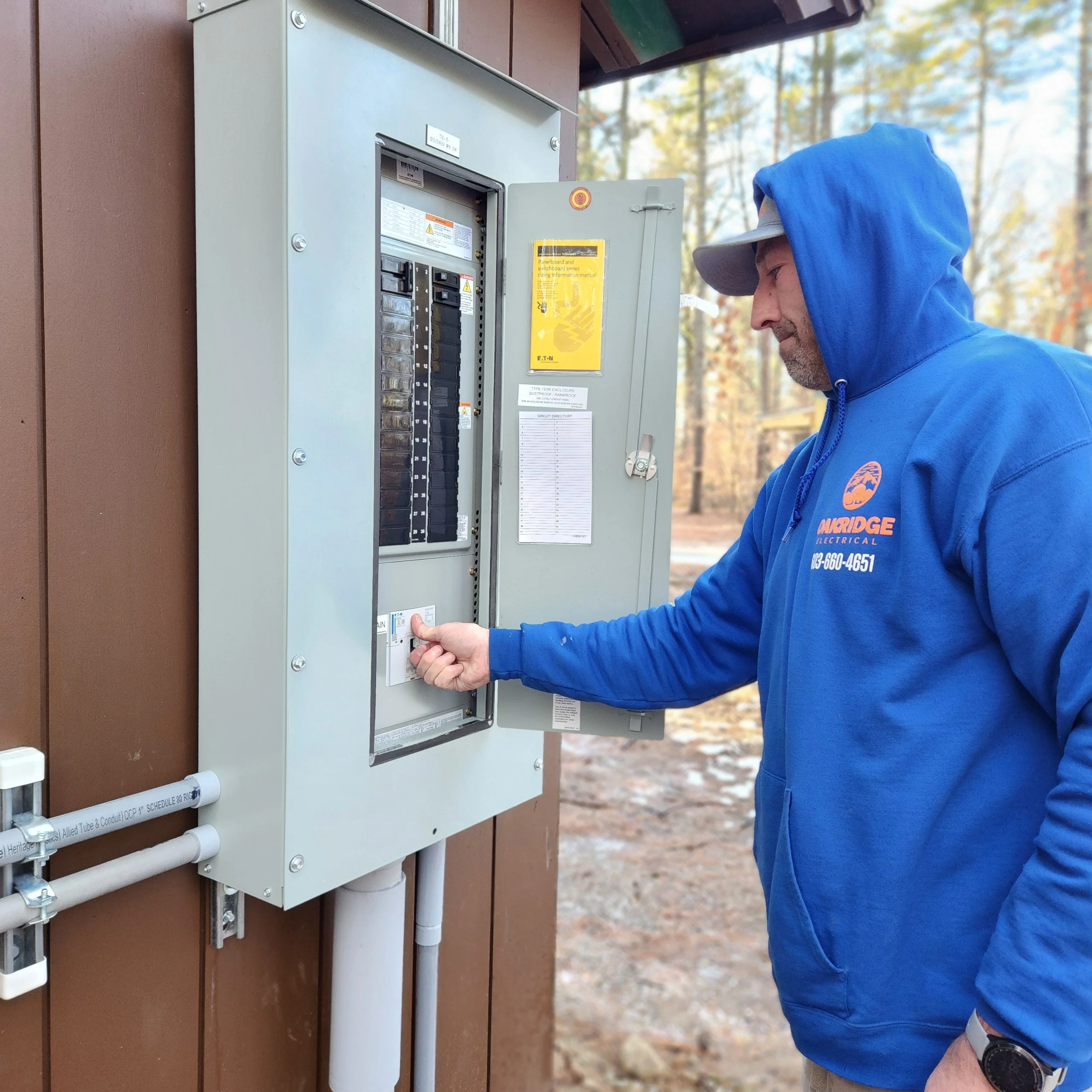 A male electrician in a blue hoodie with the logo 'Orridge Electrical' is interacting with an electrical panel outdoors on a building's exterior wall, with trees in the background.
