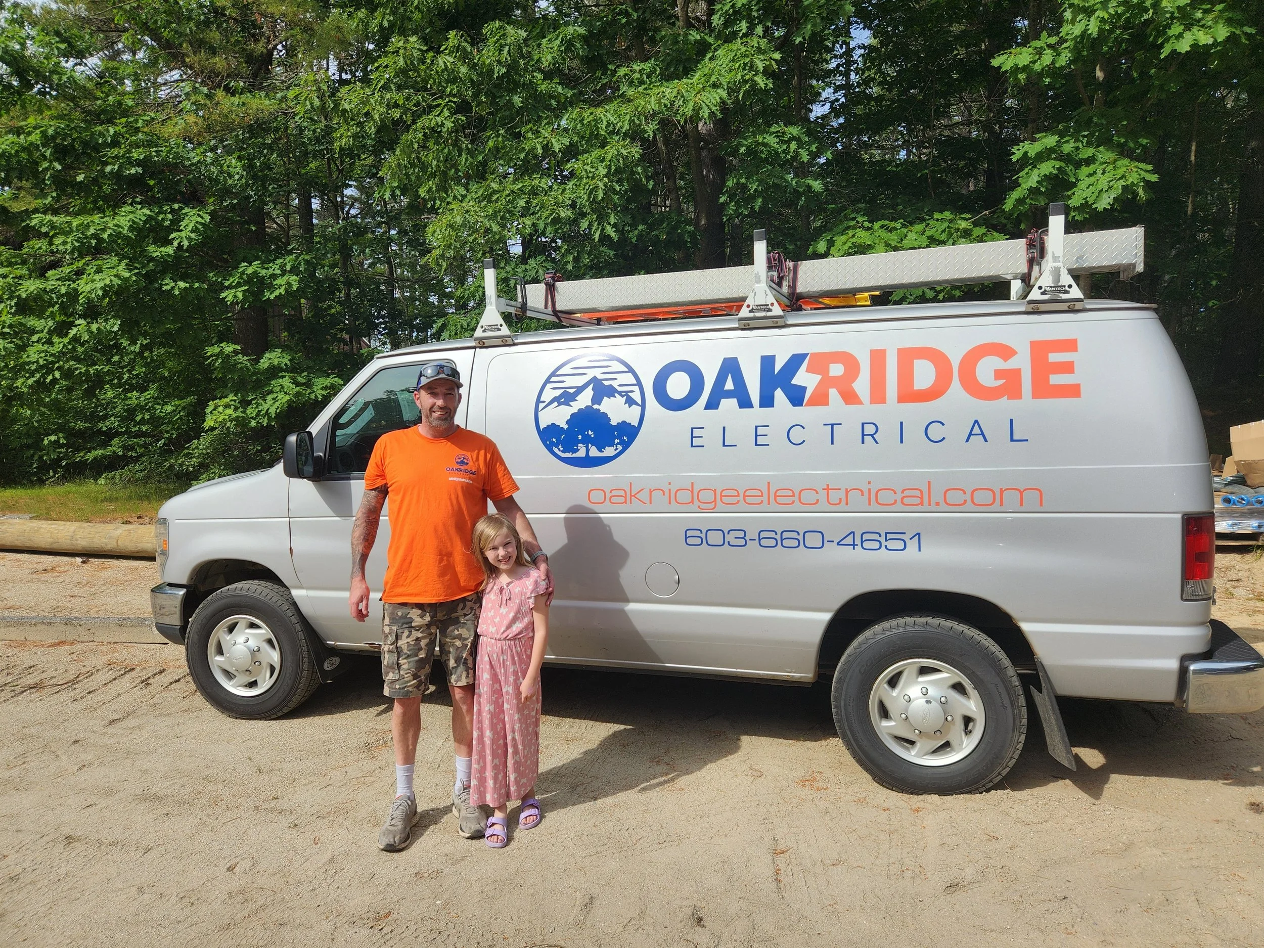 A man and a young girl standing in front of a white van with the logo and contact information of Oak Ridge Electrical. The man is wearing an orange t-shirt and camouflage shorts, and the girl is wearing a pink dress. The van is parked outdoors on a d