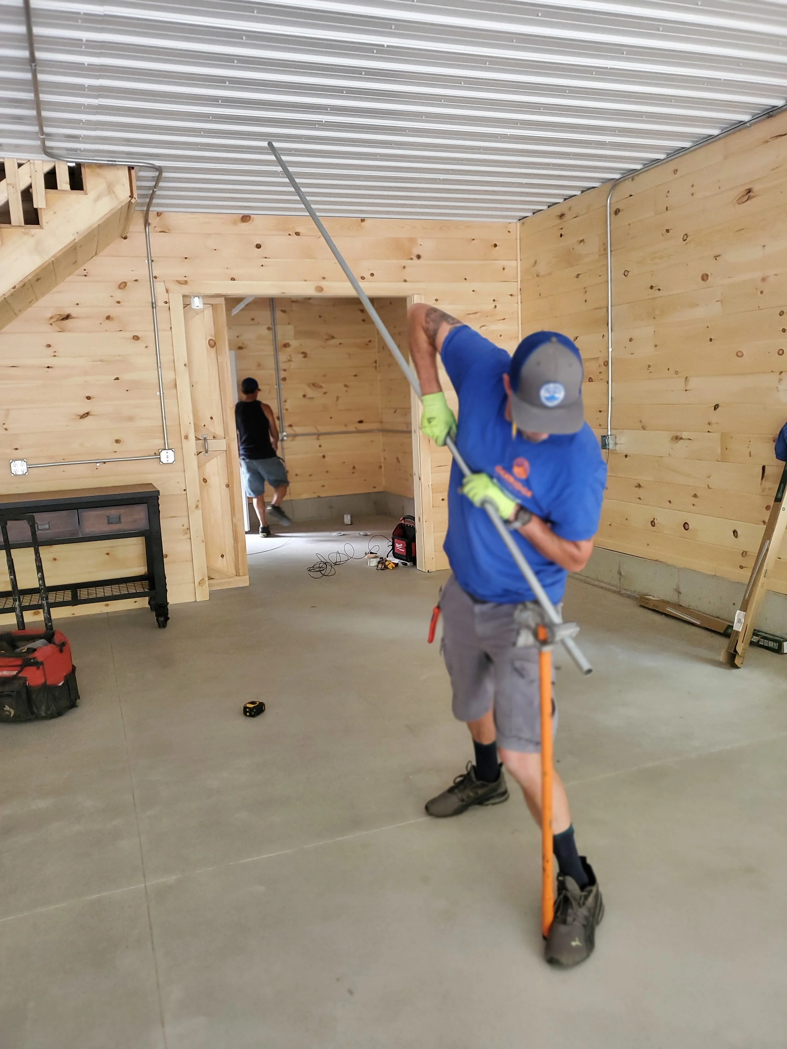 Worker in a blue shirt and gray shorts using a long-handled tool to trim or adjust the ceiling of an unfinished room with wooden walls and concrete floor.