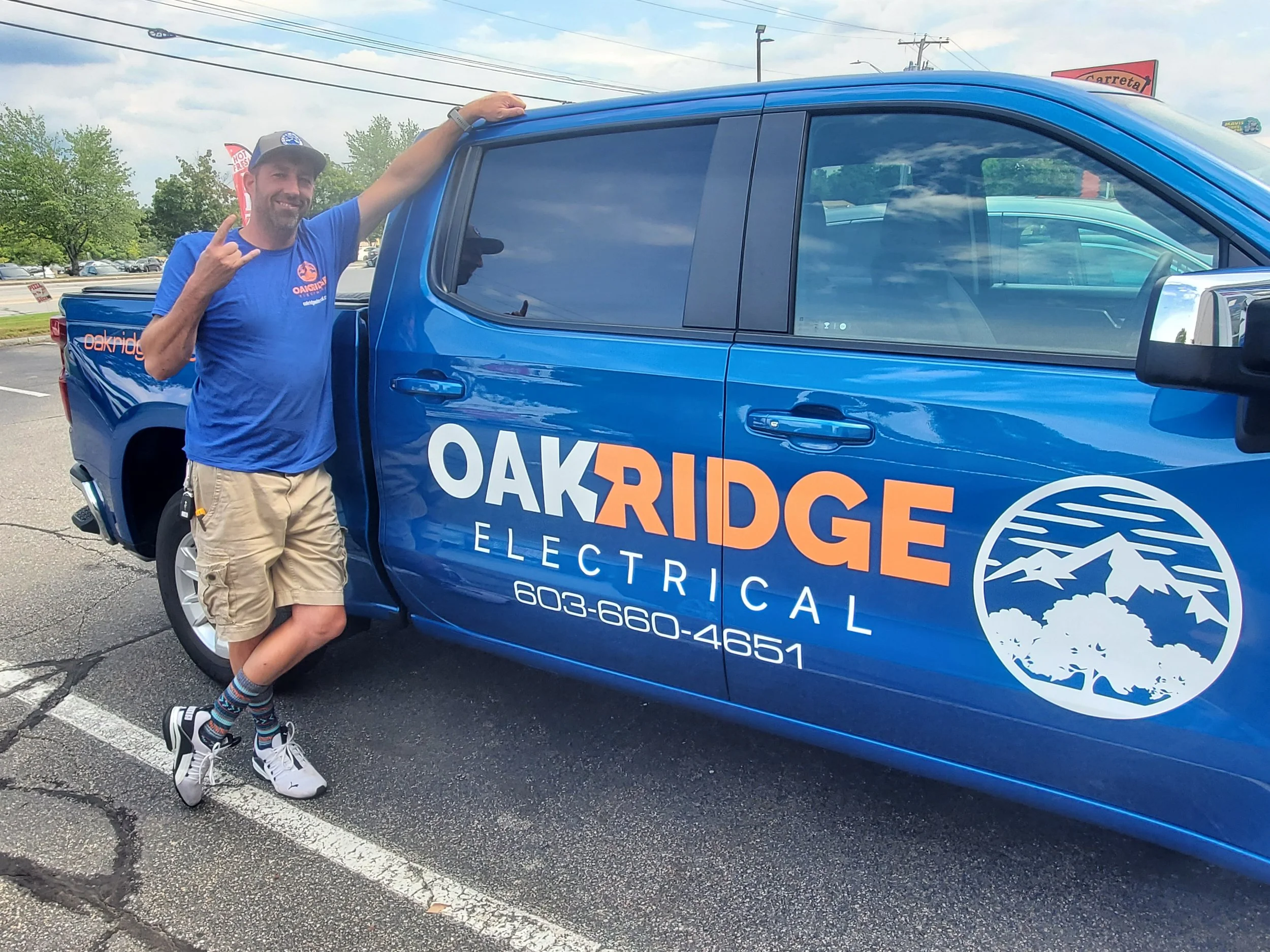 Man standing next to a blue Oak Ridge Electrical company truck in a parking lot, smiling, wearing a blue T-shirt