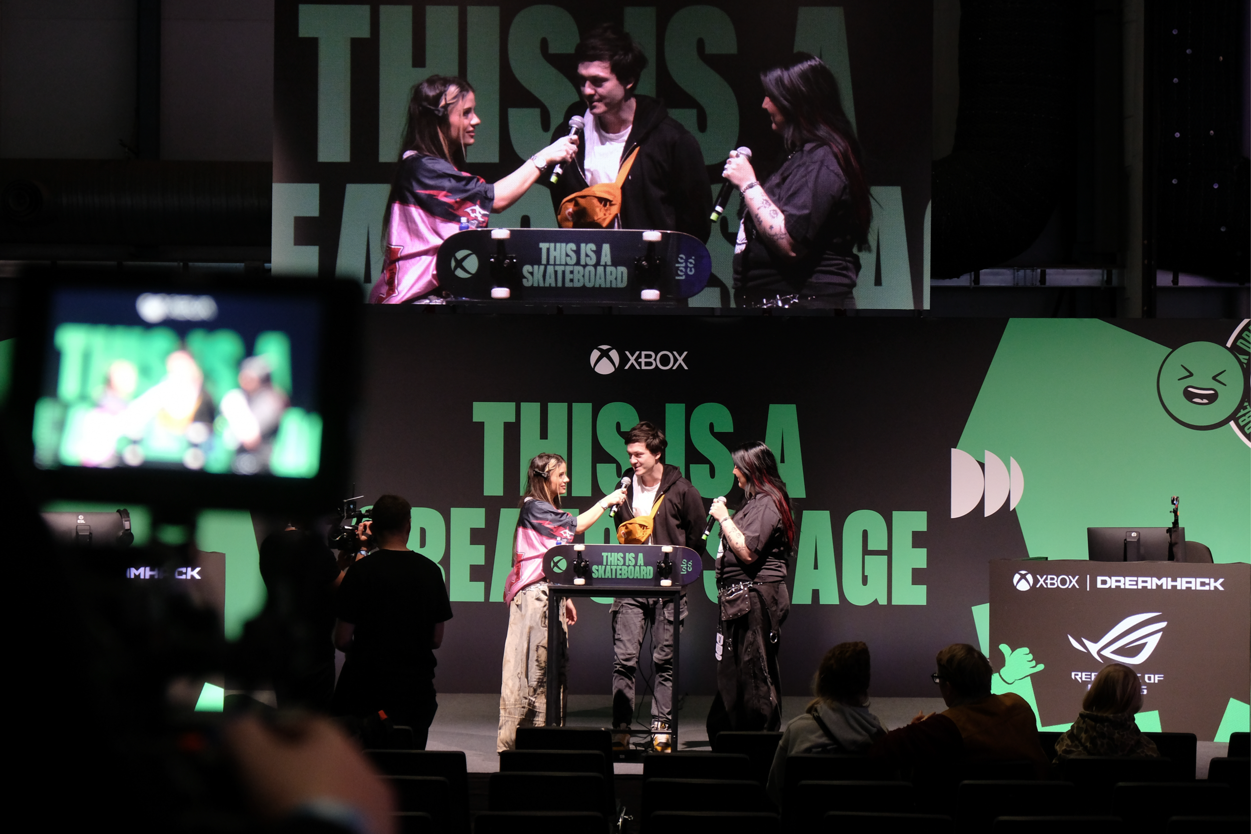 Three people are on stage at a gaming event, engaging in an interview with microphones. Behind them is a large screen with the text "THIS IS A SKATEBOARD." The event appears to be related to Xbox and DreamHack, as indicated by banners and logos.