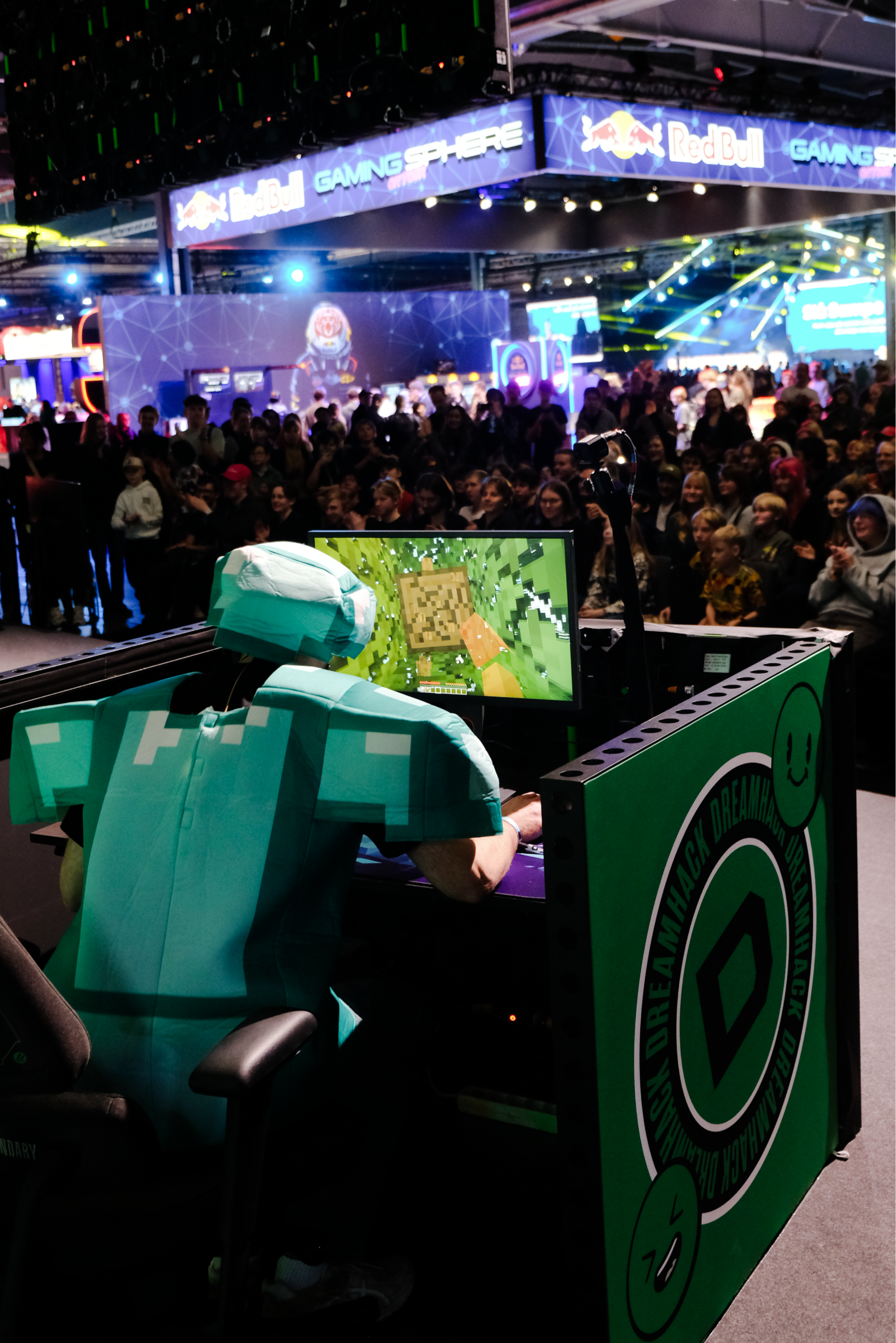 Person dressed in a Minecraft-themed cosplay with a pixelated green outfit, sitting at a gaming station in a crowded gaming event or convention.