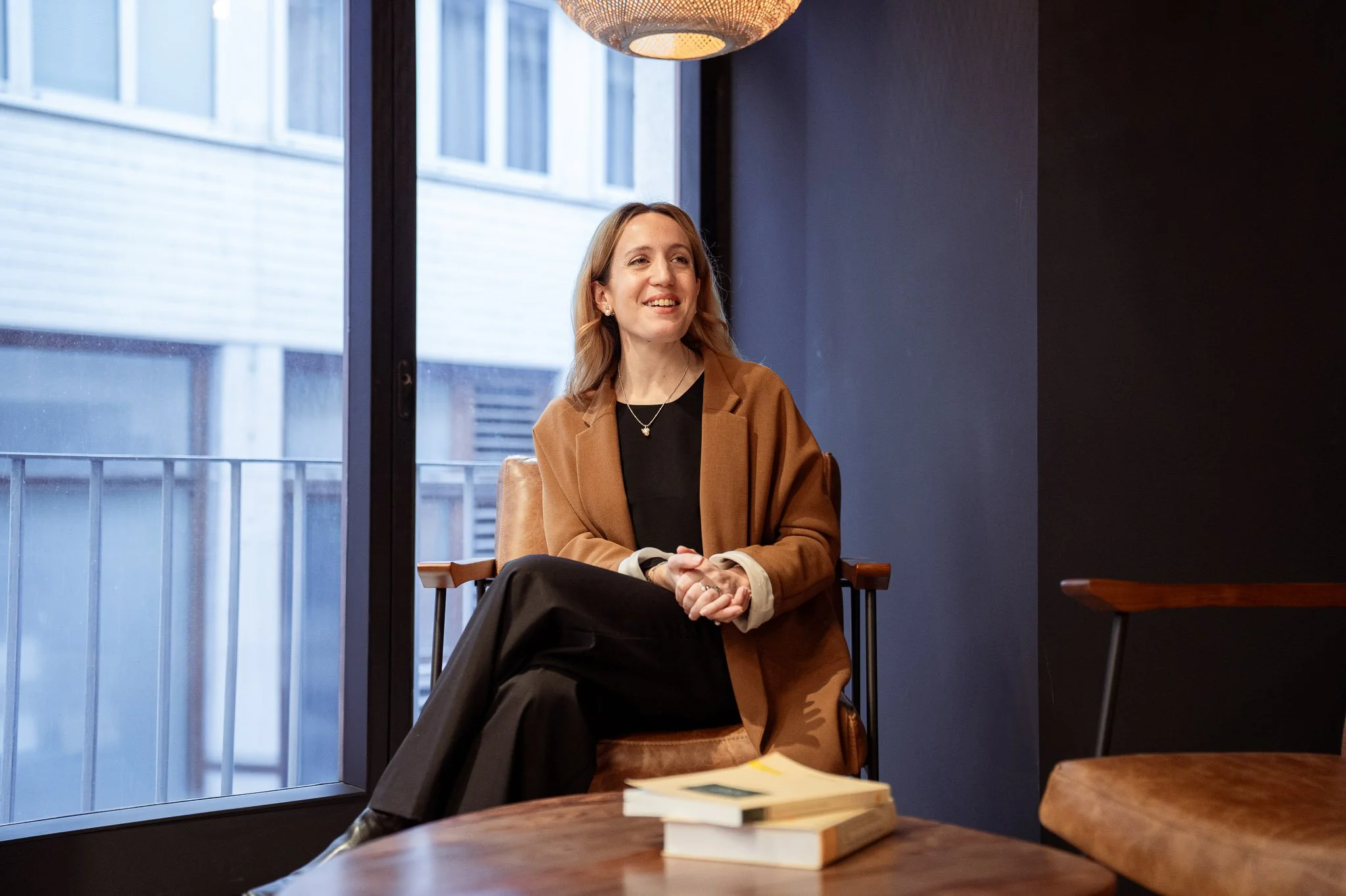 A woman with blonde hair, wearing a brown blazer over a black top, sitting in a room with a large window. She is smiling, with books on a table in front of her.