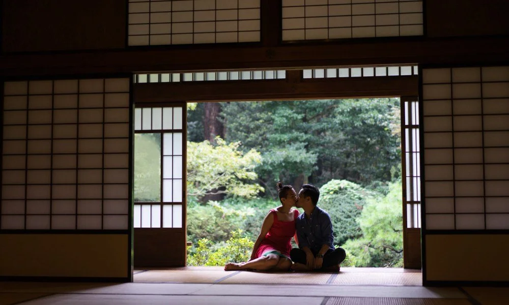 A couple sitting on tatami mats inside a traditional Japanese-style room, sharing a kiss with a view of a lush garden outside.