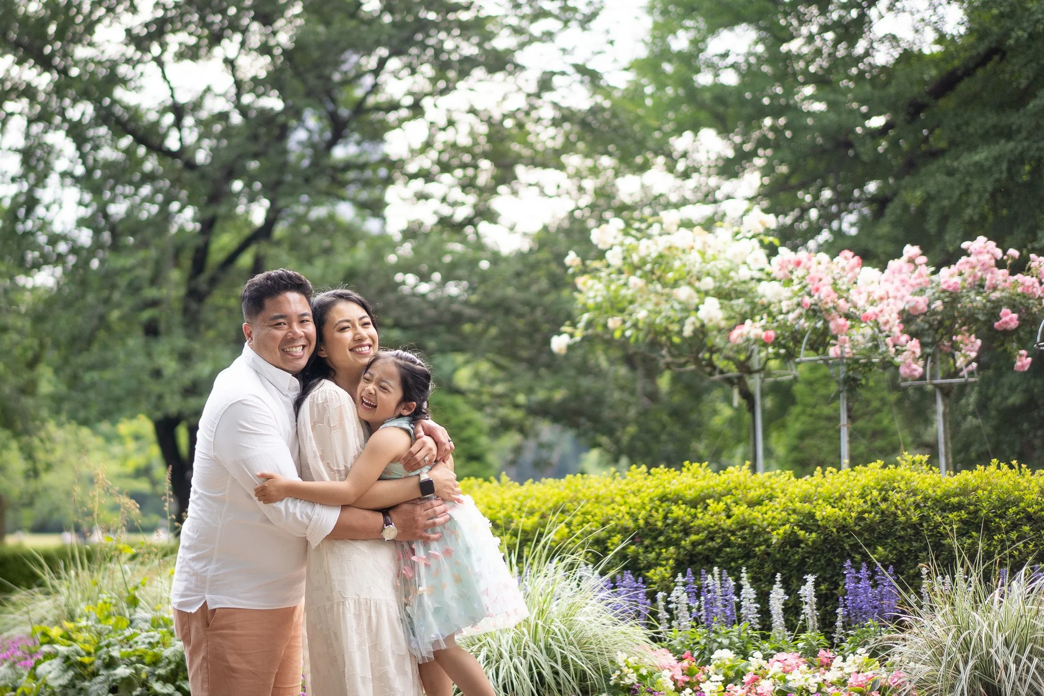 A happy family of three hugging in a lush garden with flowering plants and trees.