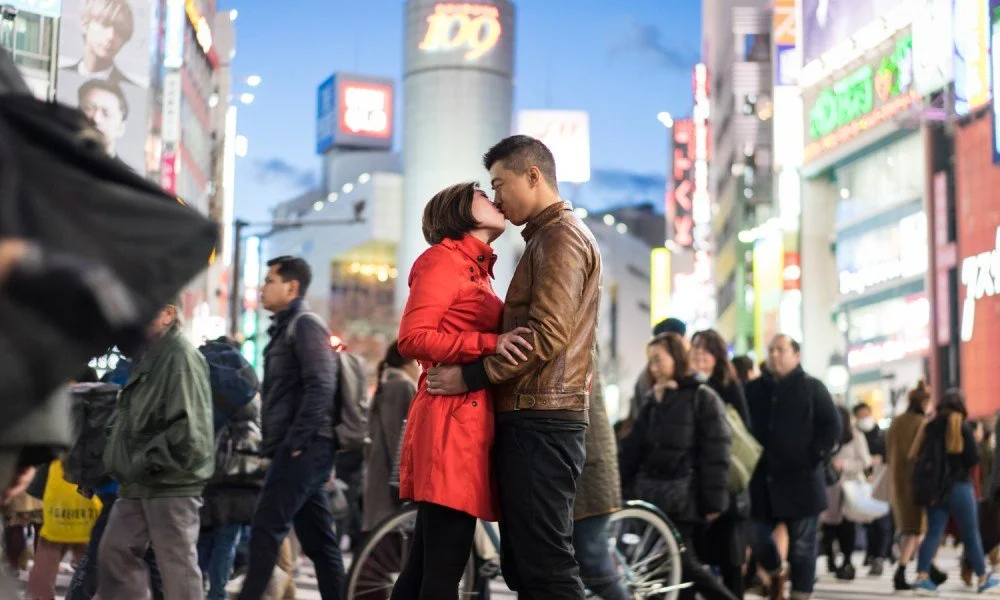 A couple kissing on a busy city street at night, surrounded by pedestrians and colorful illuminated signs.