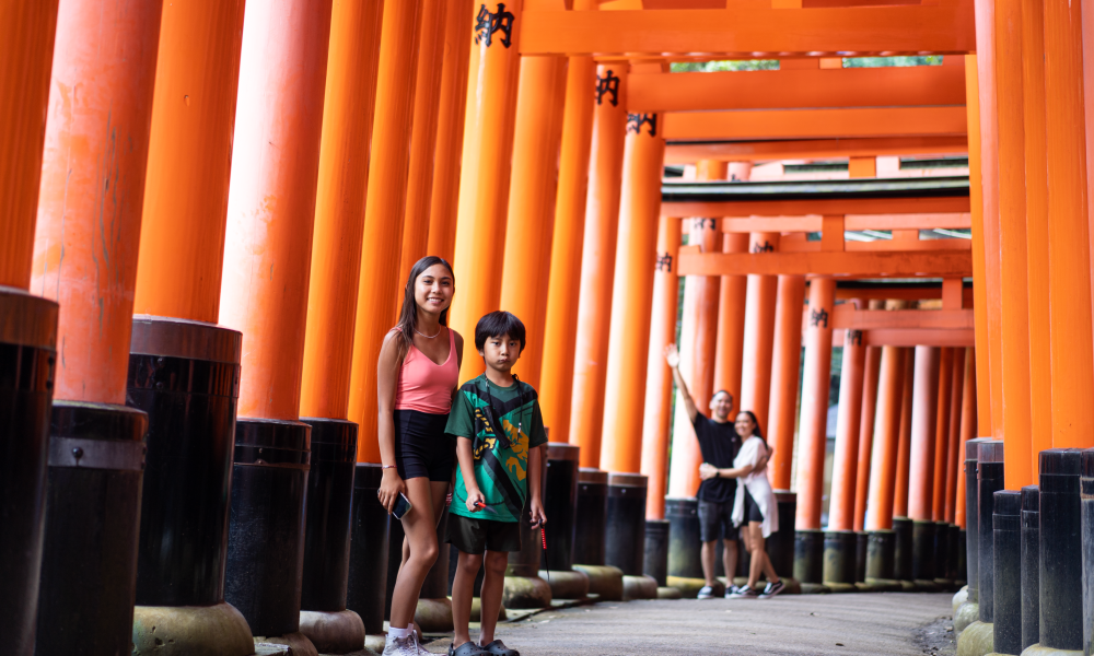 Two children, a girl and a boy, pose under a series of red torii gates at Fushimi Inari Shrine in Japan, with two adults in the background taking a photo.