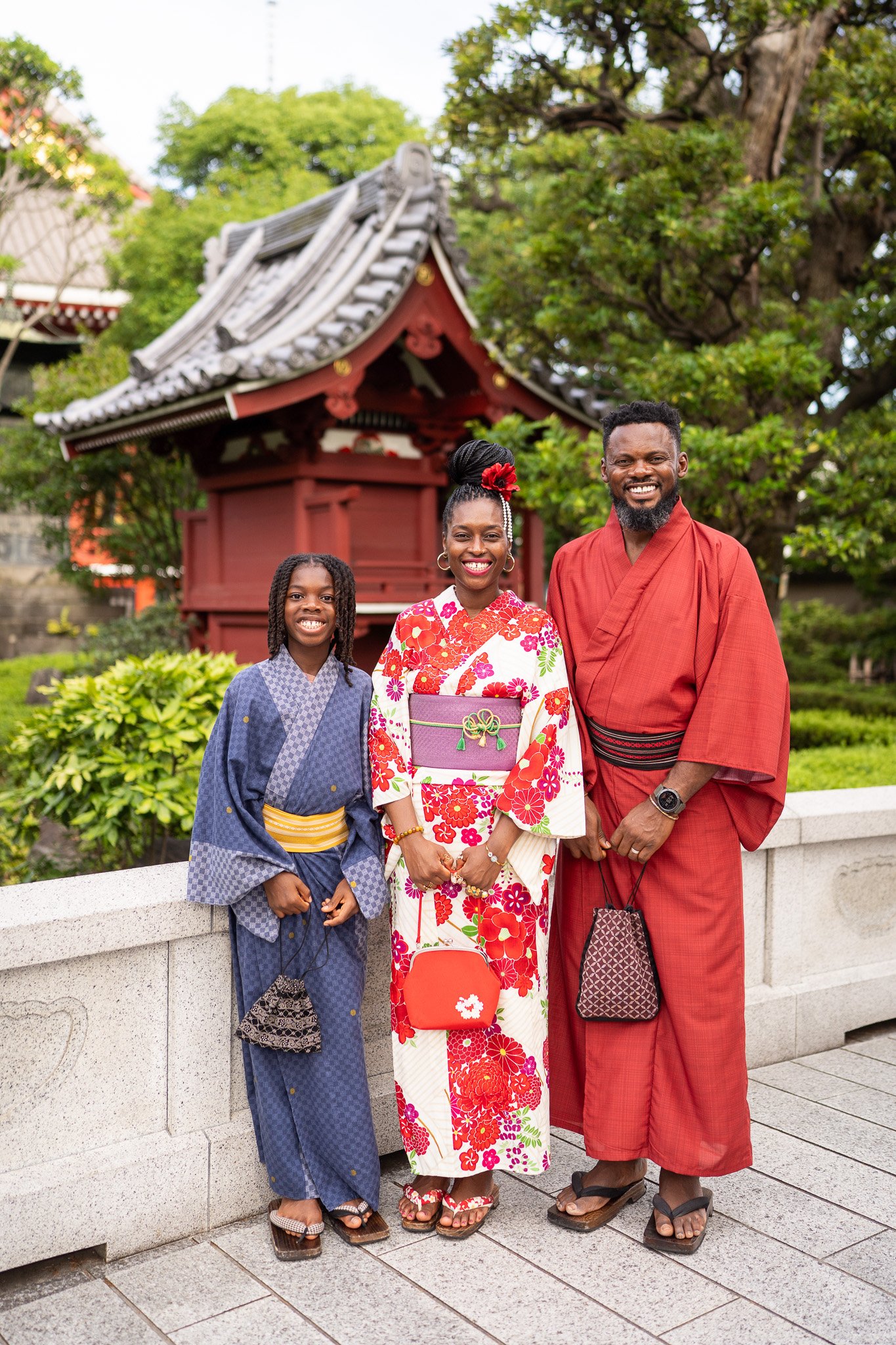 A family of three dressed in traditional Japanese kimonos, standing outdoors in front of a Japanese-style structure and lush green trees, smiling.