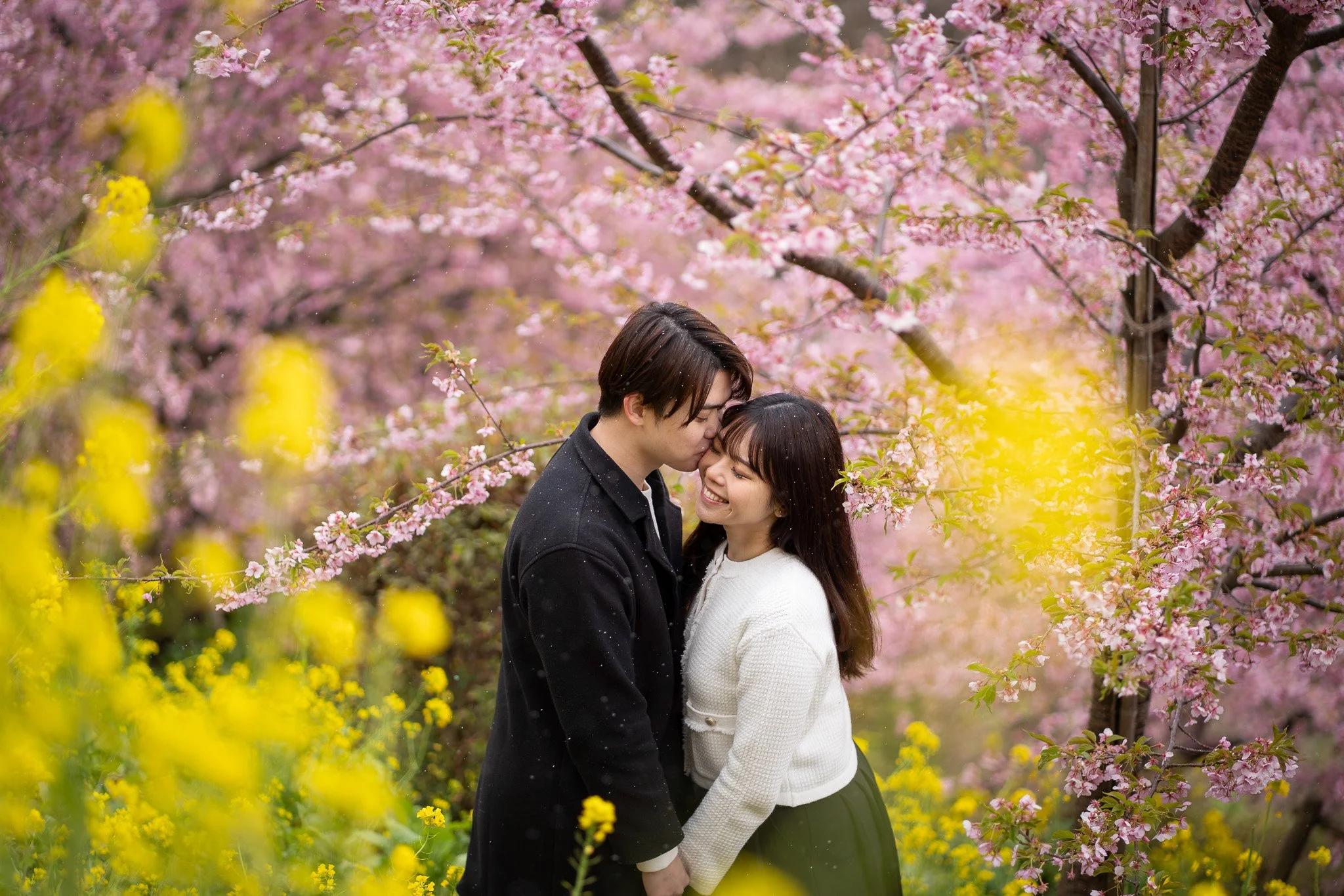 A couple smiling and embracing in a garden with pink cherry blossoms and yellow flowers.