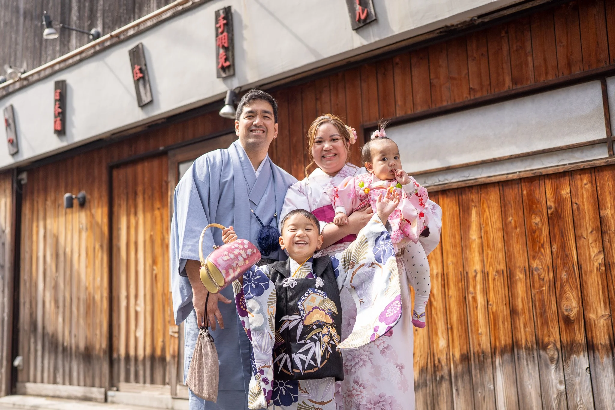 Family of four dressed in traditional Japanese clothing, smiling outside a wooden building with Japanese signs.