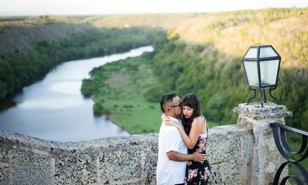 A couple embraces on a stone bridge overlooking a winding river and lush green landscape.
