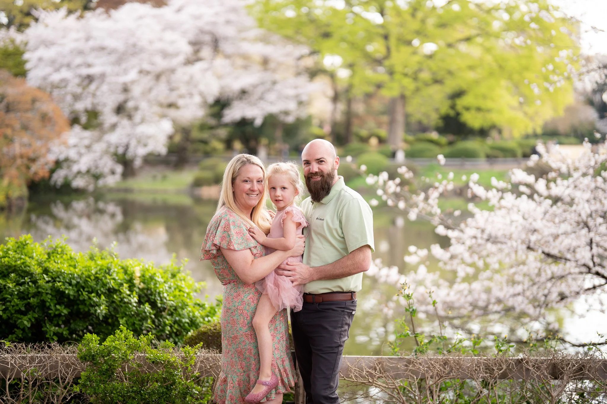 A family of three, consisting of a woman, a man, and a young girl, smiling and standing near a pond with blooming cherry trees and greenery in the background.