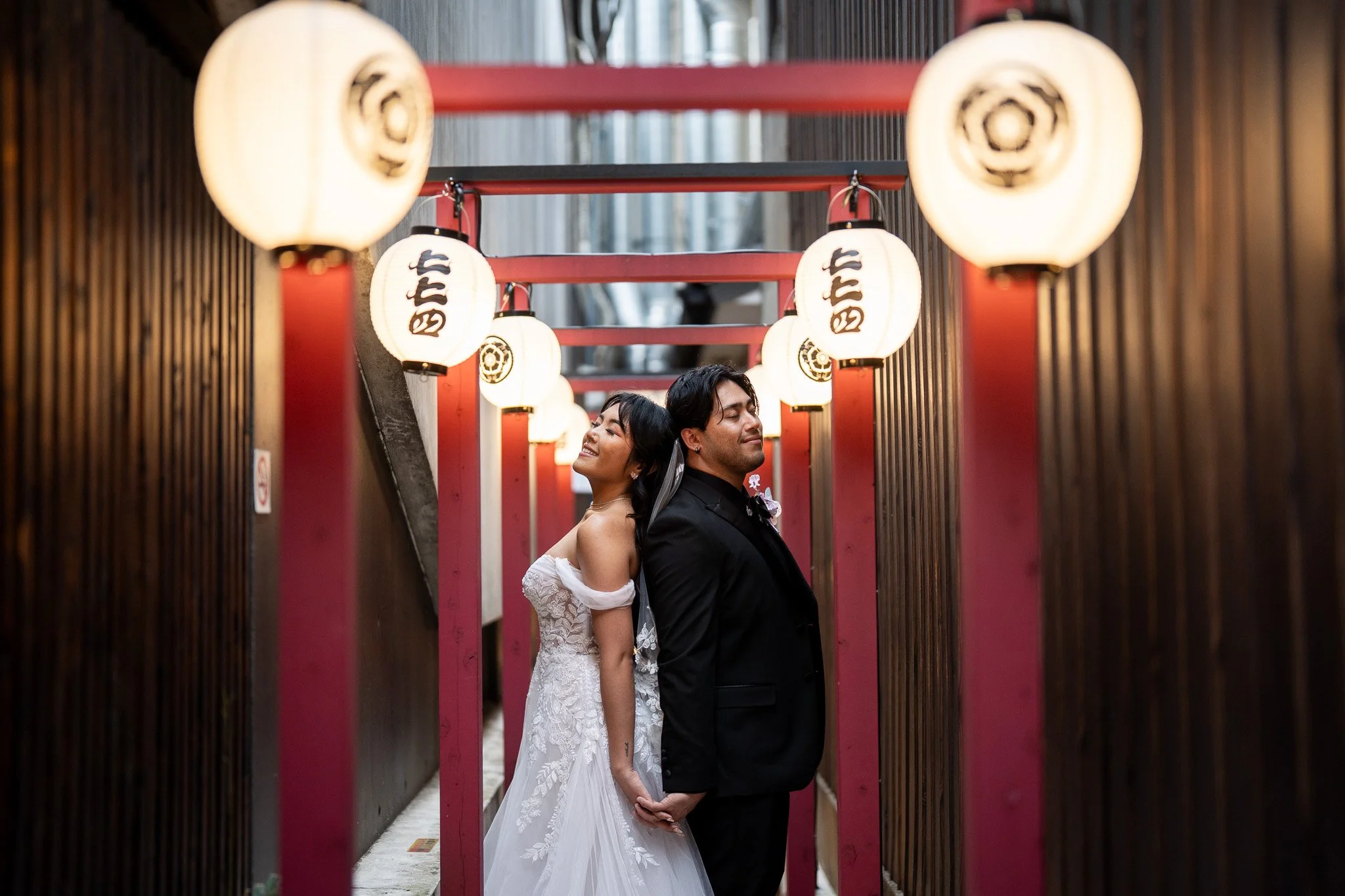 A newlywed couple holding hands and standing back-to-back near illuminated paper lanterns in a traditional Japanese setting.