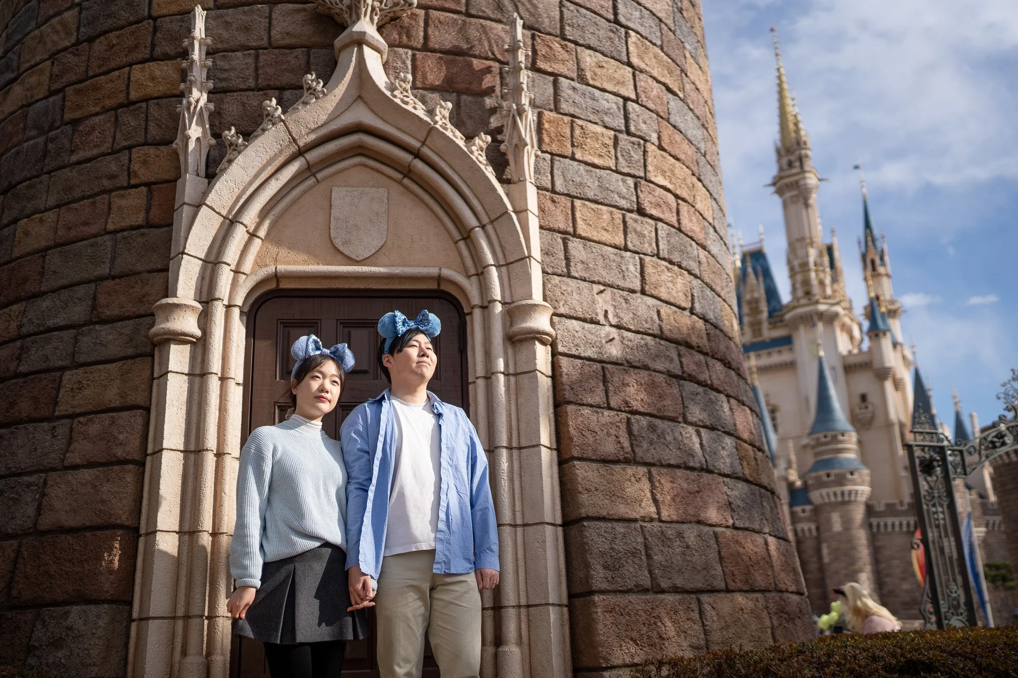 A young couple stands in front of Sleeping Beauty Castle at Disneyland, wearing matching blue Mickey Mouse ear headbands, with the castle's stone wall and turrets visible in the background.