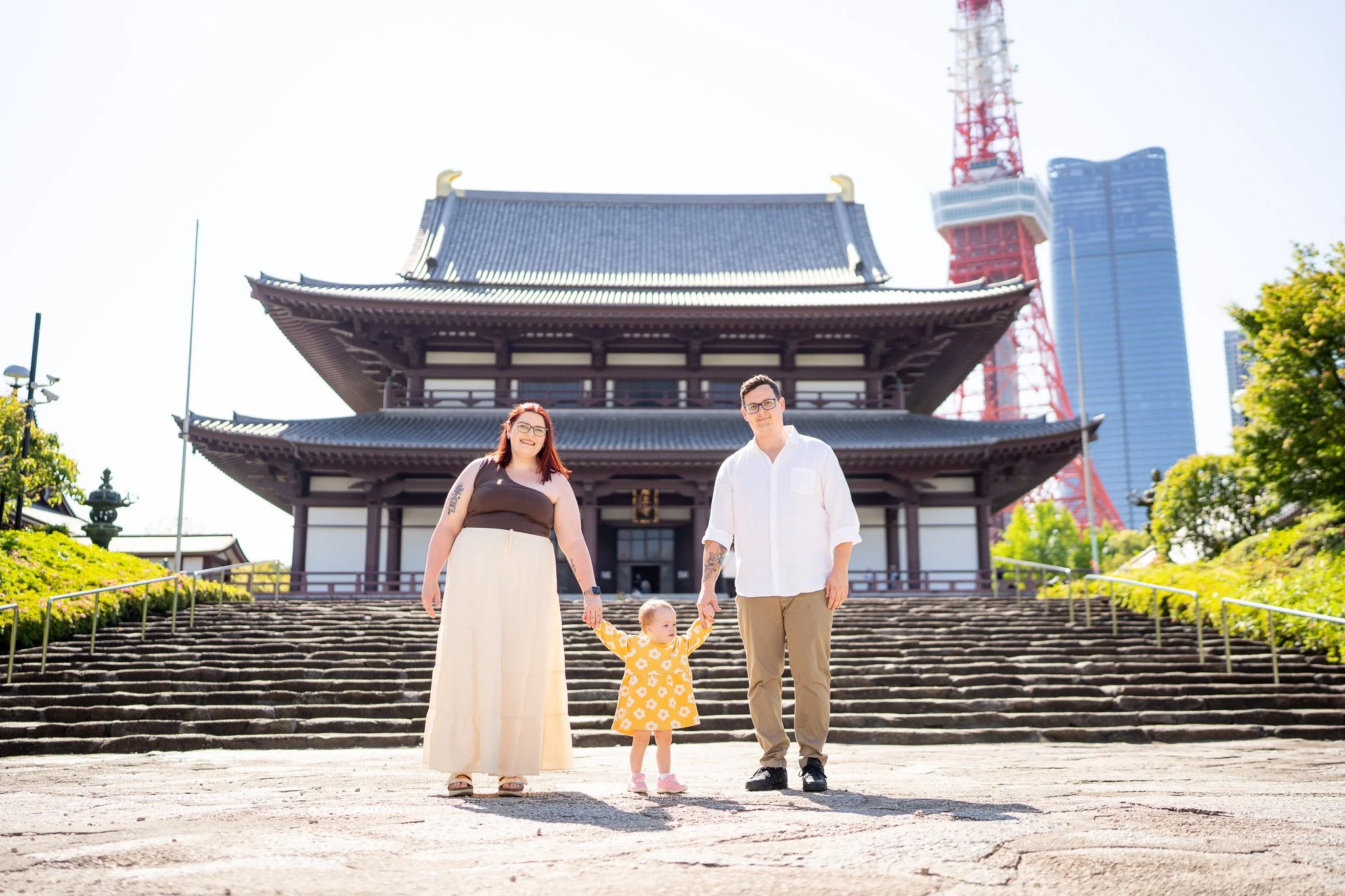 A family of three standing on steps in front of a traditional Japanese temple, with Tokyo Tower and skyscrapers in the background, during daytime.