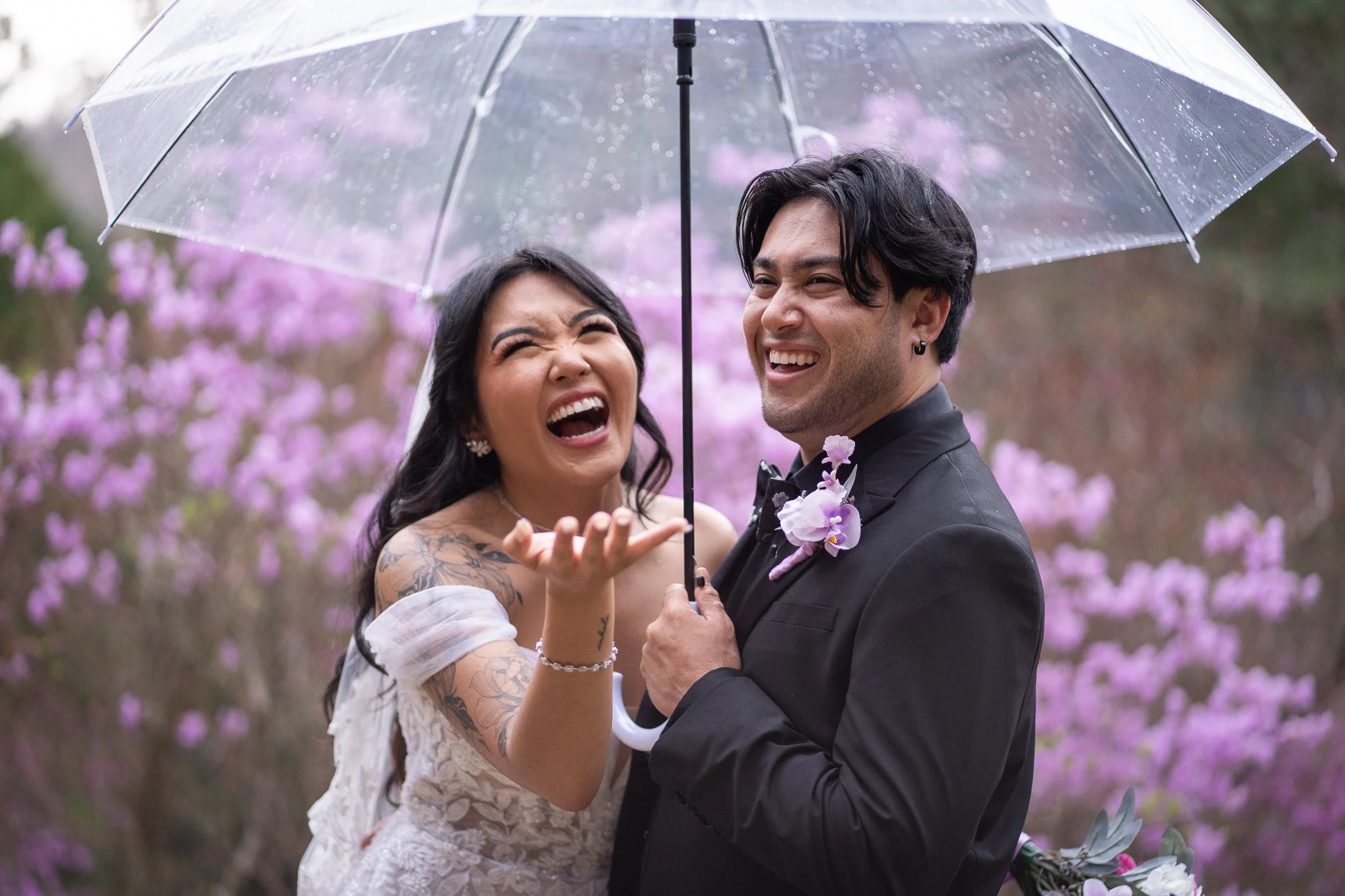 A joyful couple in wedding attire sharing a moment under an umbrella in a garden with pink blooming trees.