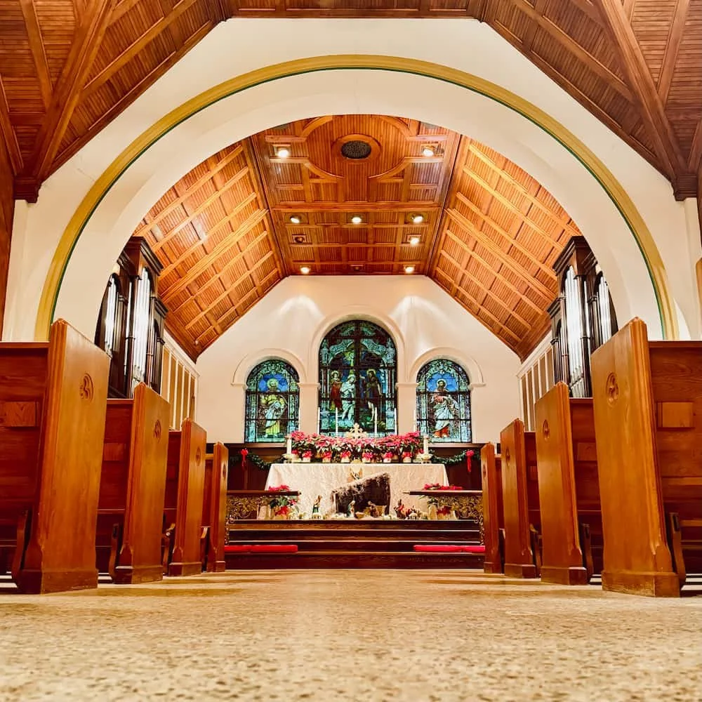 Low-angle view down the center aisle of a small church sanctuary, with wooden pews on both sides, a warm-toned vaulted wood ceiling and large arch overhead, and an altar decorated with red poinsettias in front of three stained-glass windows.