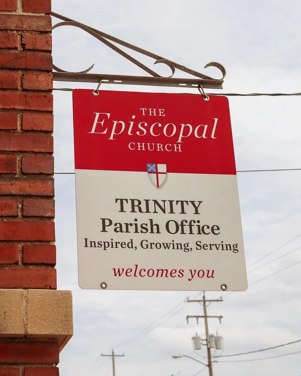 Sign for The Episcopal Church Trinity Parish Office on a brick building, with power lines and a cloudy sky in the background.