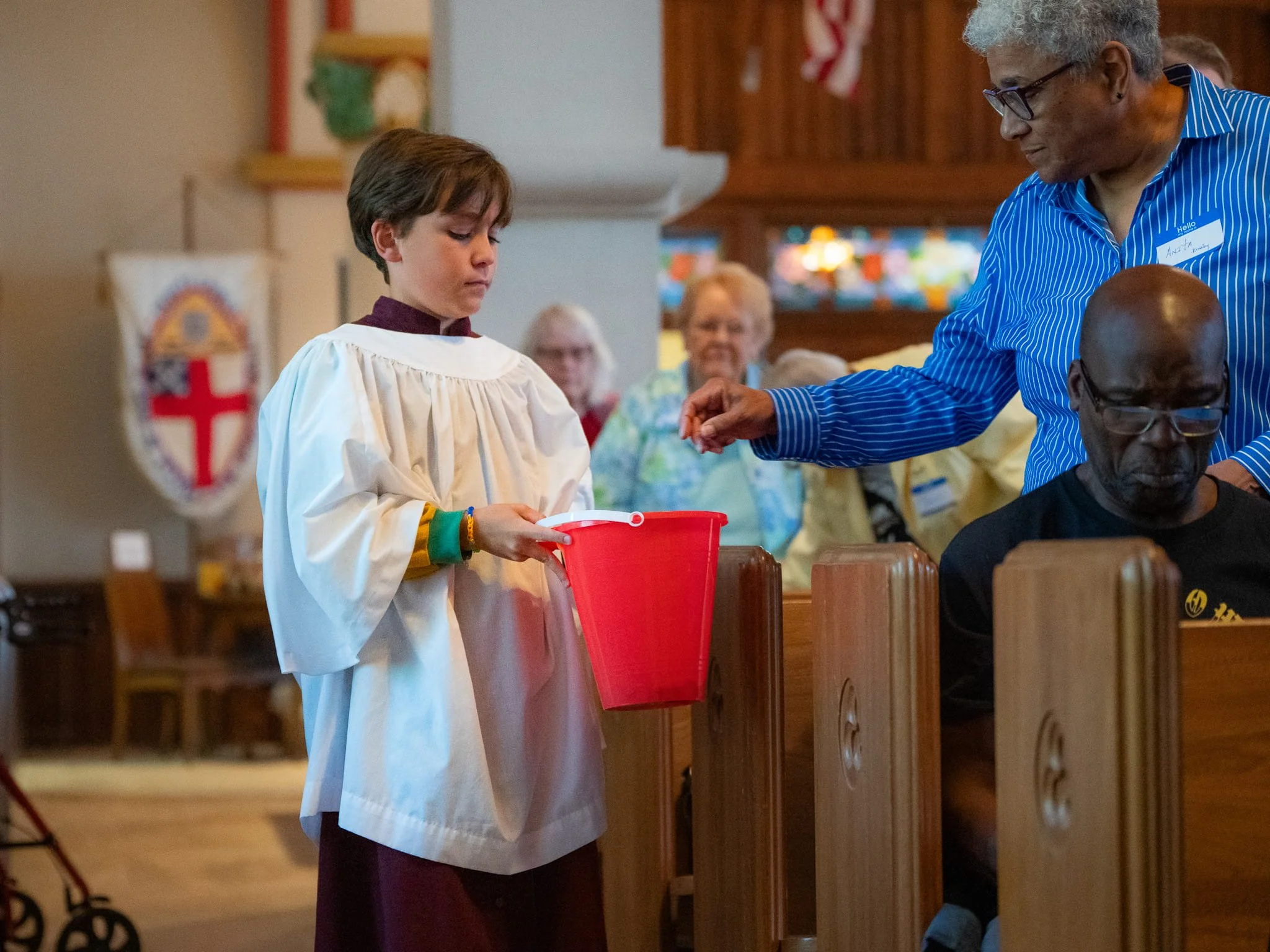 A young boy in a choir robe placing an offering into a red collection bucket held by a woman at a church service.