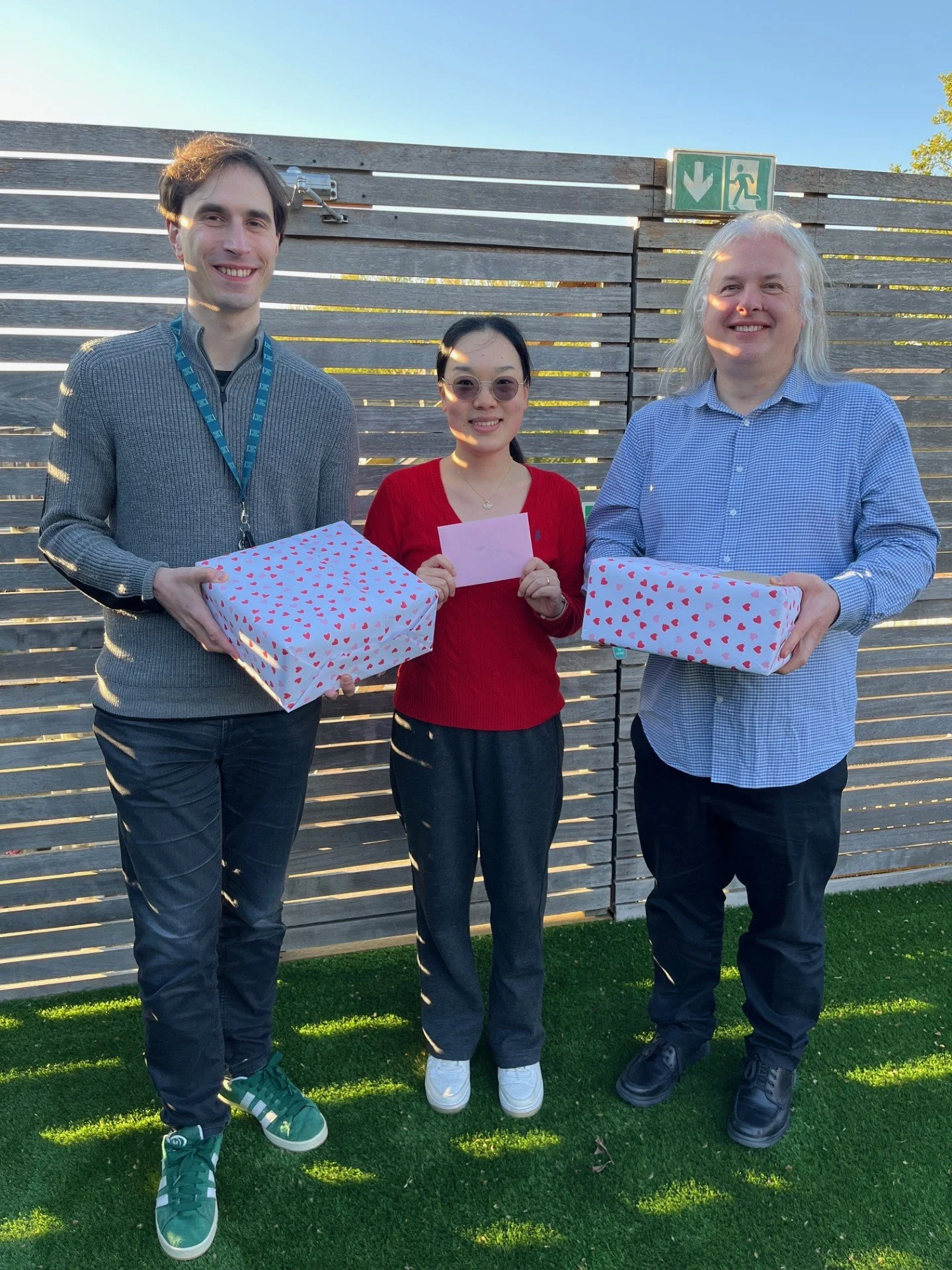 Three people standing outdoors in front of a wooden fence, holding gift boxes with heart patterns, smiling, during daytime.