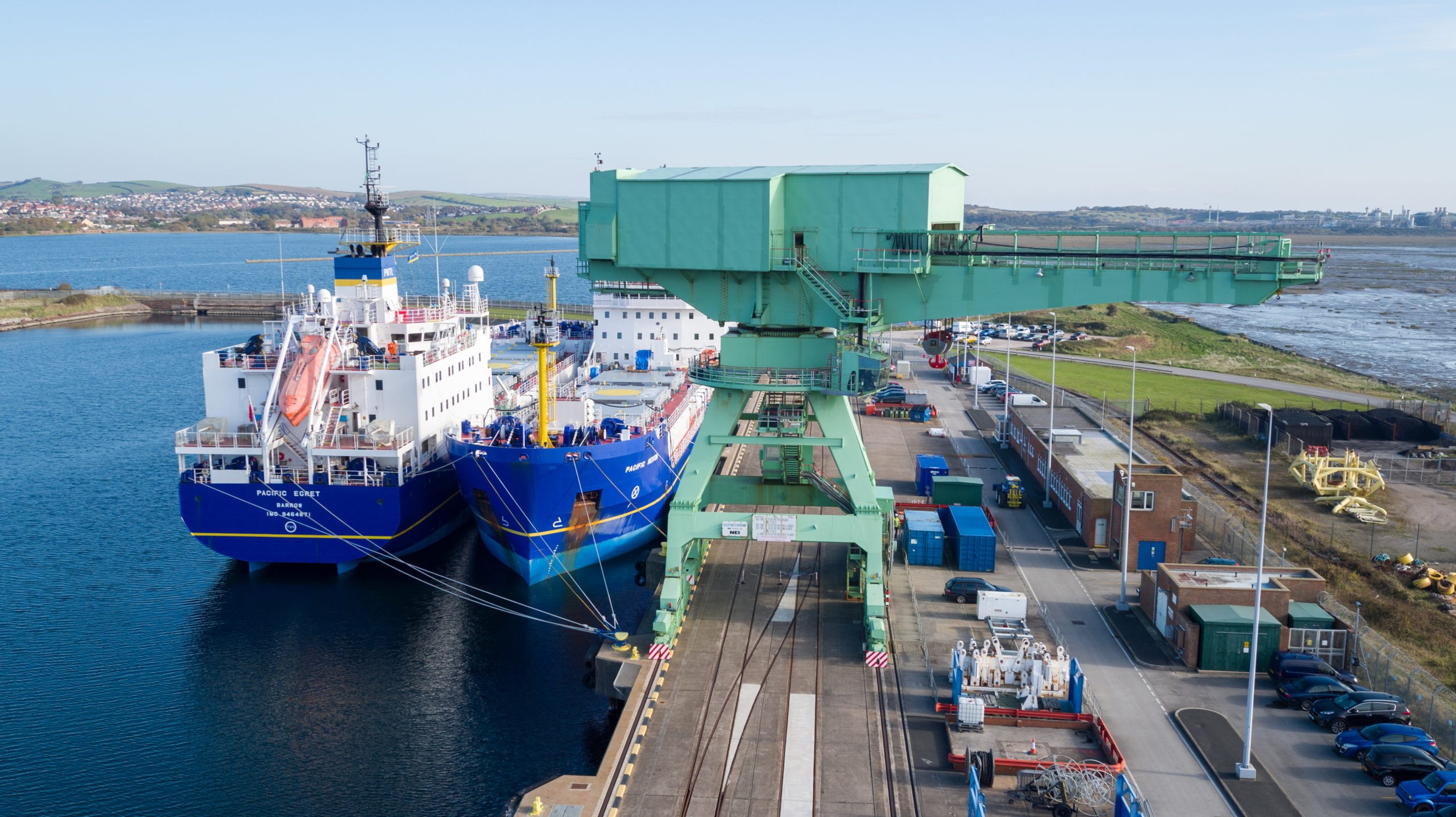 Large green crane loading or unloading cargo ship called Pacific Egret in a dockyard with parked vehicles and industrial equipment.