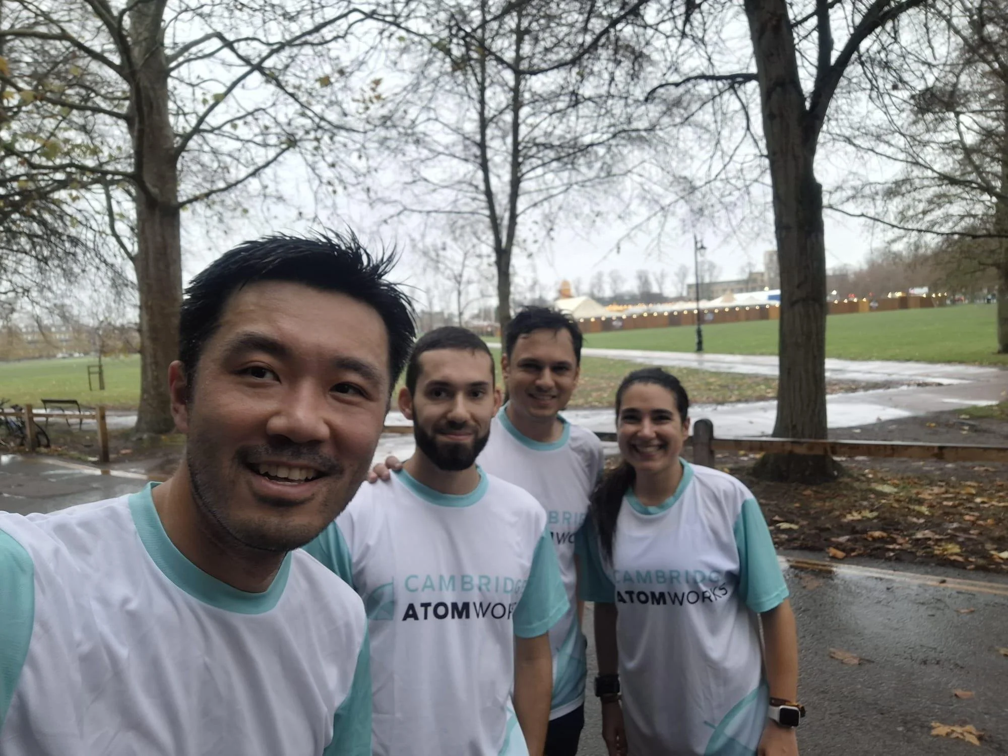 Four people in athletic shirts taking a group selfie outdoors in a park with leafless trees and a cloudy sky.