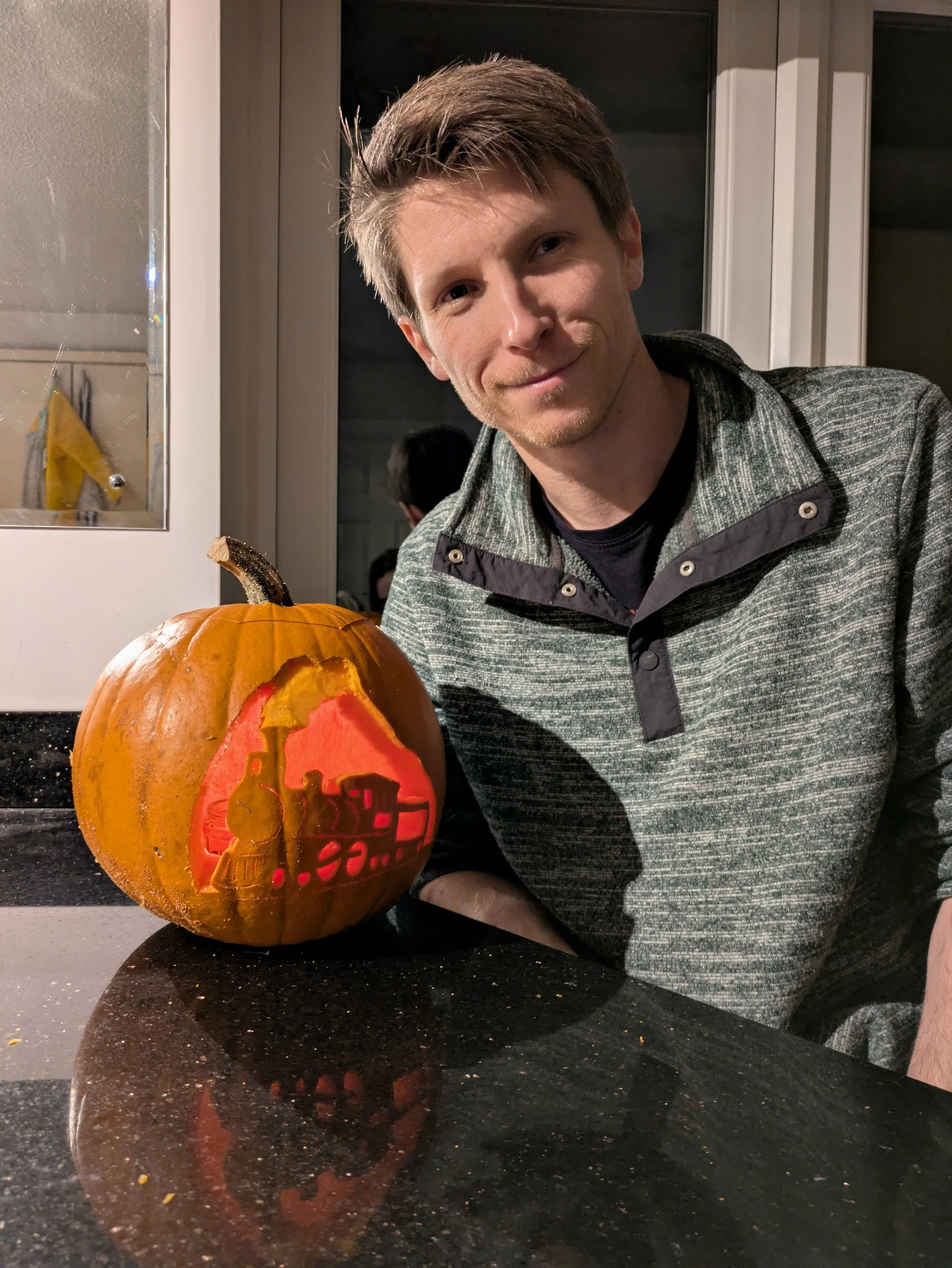 Man with short brown hair and a light beard smiling next to a carved pumpkin with a train scene on it, on a black countertop in a kitchen.