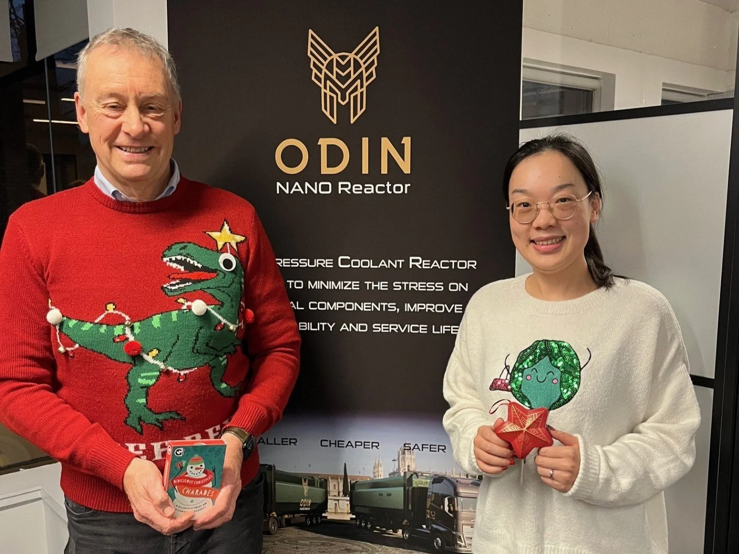 Two people standing indoors near a black backdrop with golden text and logo that reads 'ODIN NANO Reactor'. The man on the left is wearing a red Christmas sweater with a cartoon dinosaur decorated with string lights and a star, and is holding a phone with a Christmas themed case. The woman on the right is wearing a white sweater with a green cartoon character on it, holding a small plush toy or ornament.