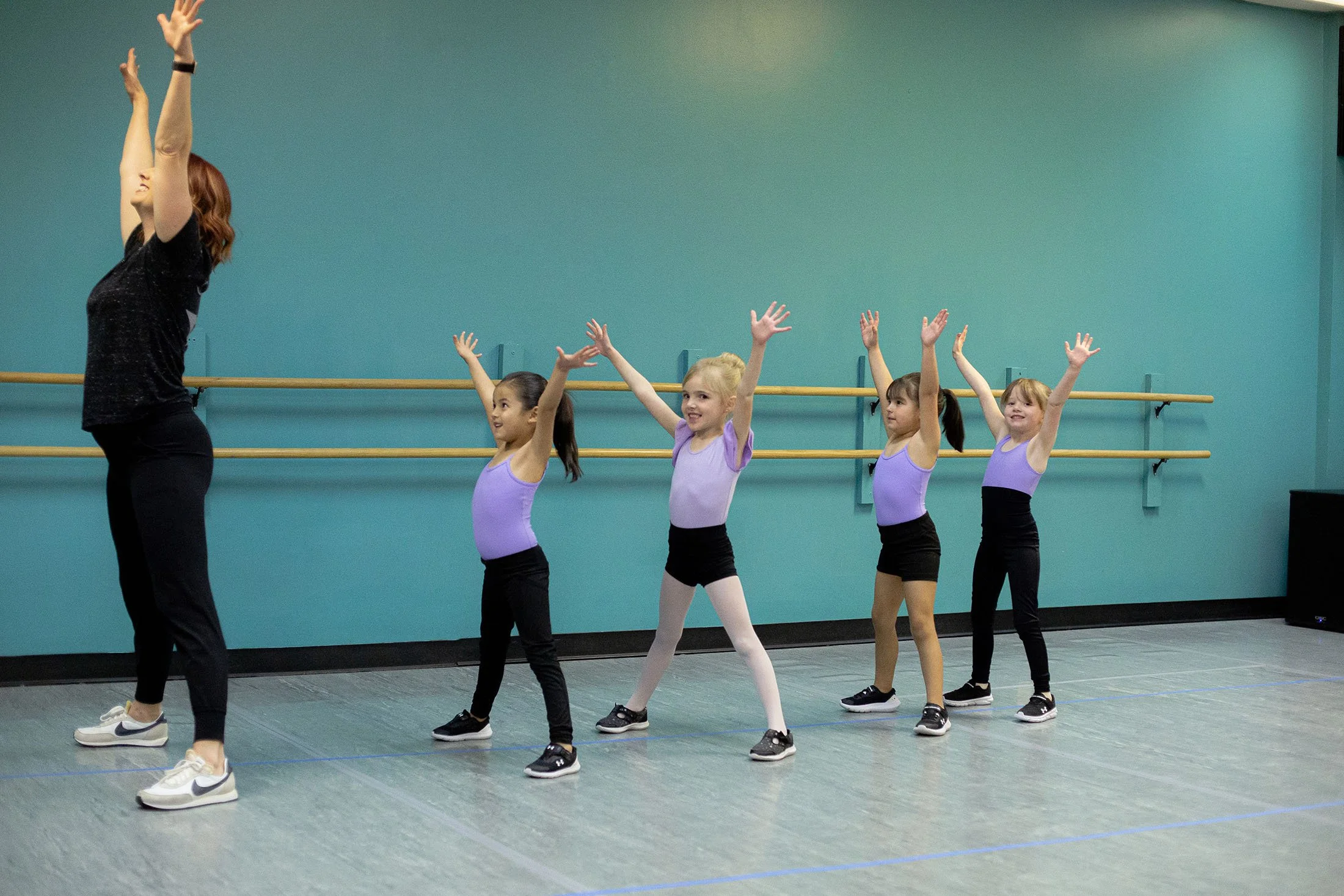 A ballet instructor leading a class of young girls in a dance studio, with all the girls raising their arms and smiling.