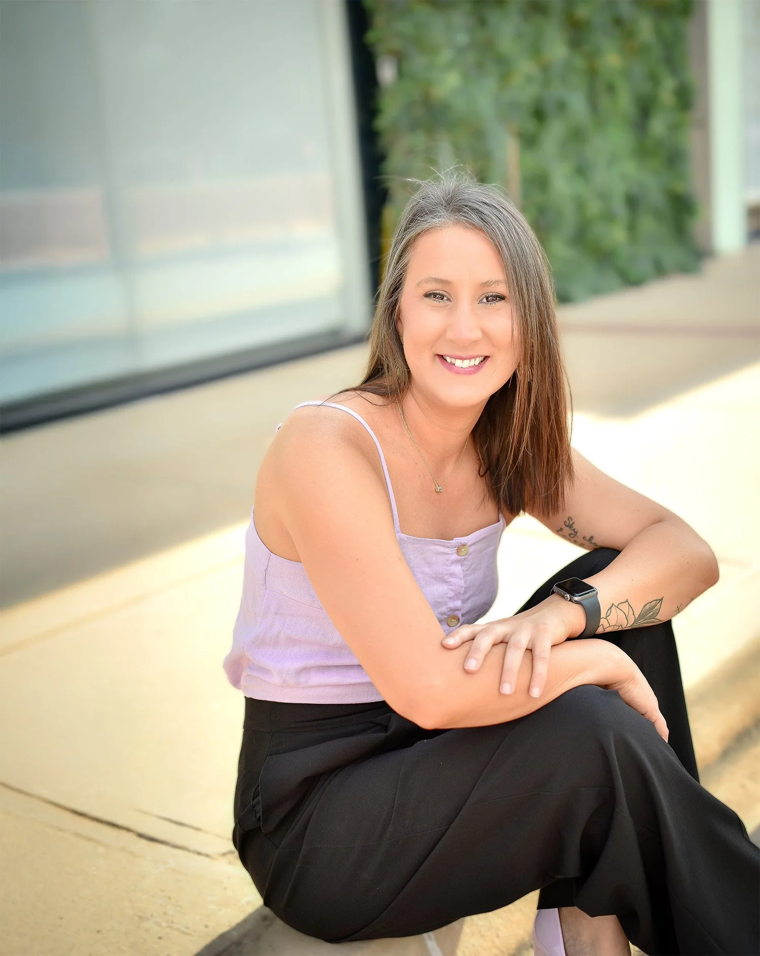 A smiling woman with long brown hair sitting outdoors on a sidewalk, wearing a lavender tank top, black pants, and a smartwatch, with a green background.