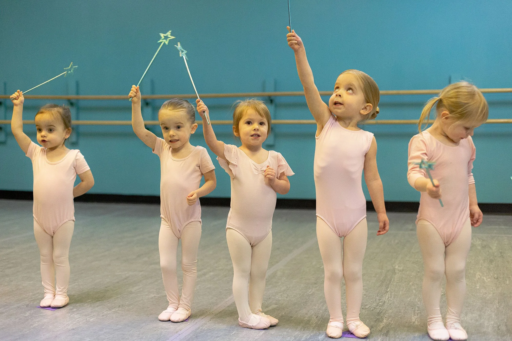 Five young girls in pink ballet leotards and tights standing on a dance studio floor, holding star-shaped magic wands with sparkling star tops, participating in a ballet class or performance.