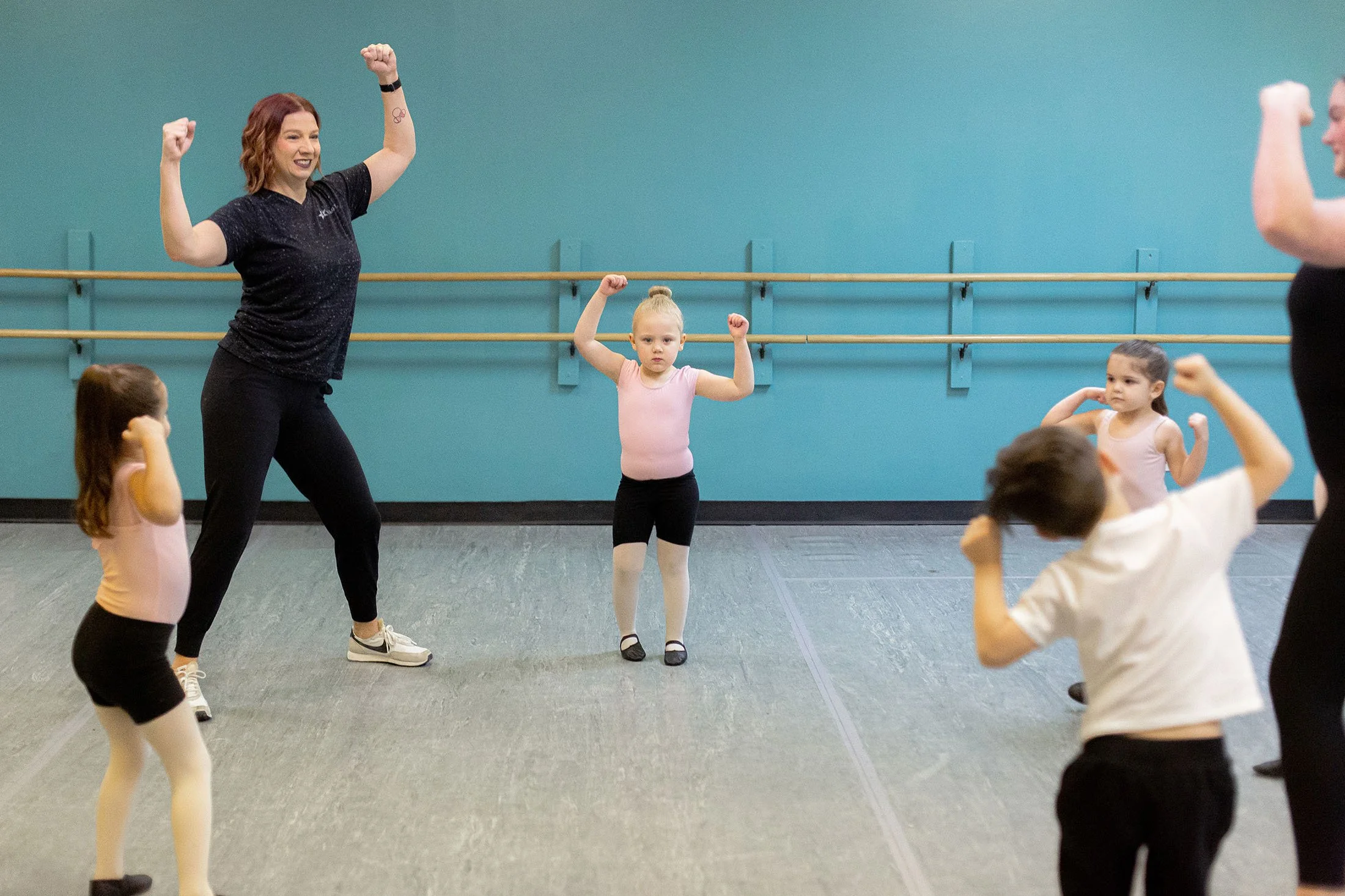 Children in ballet class with instructor, stretching and raising arms in a dance studio with blue walls and wood ballet barres.
