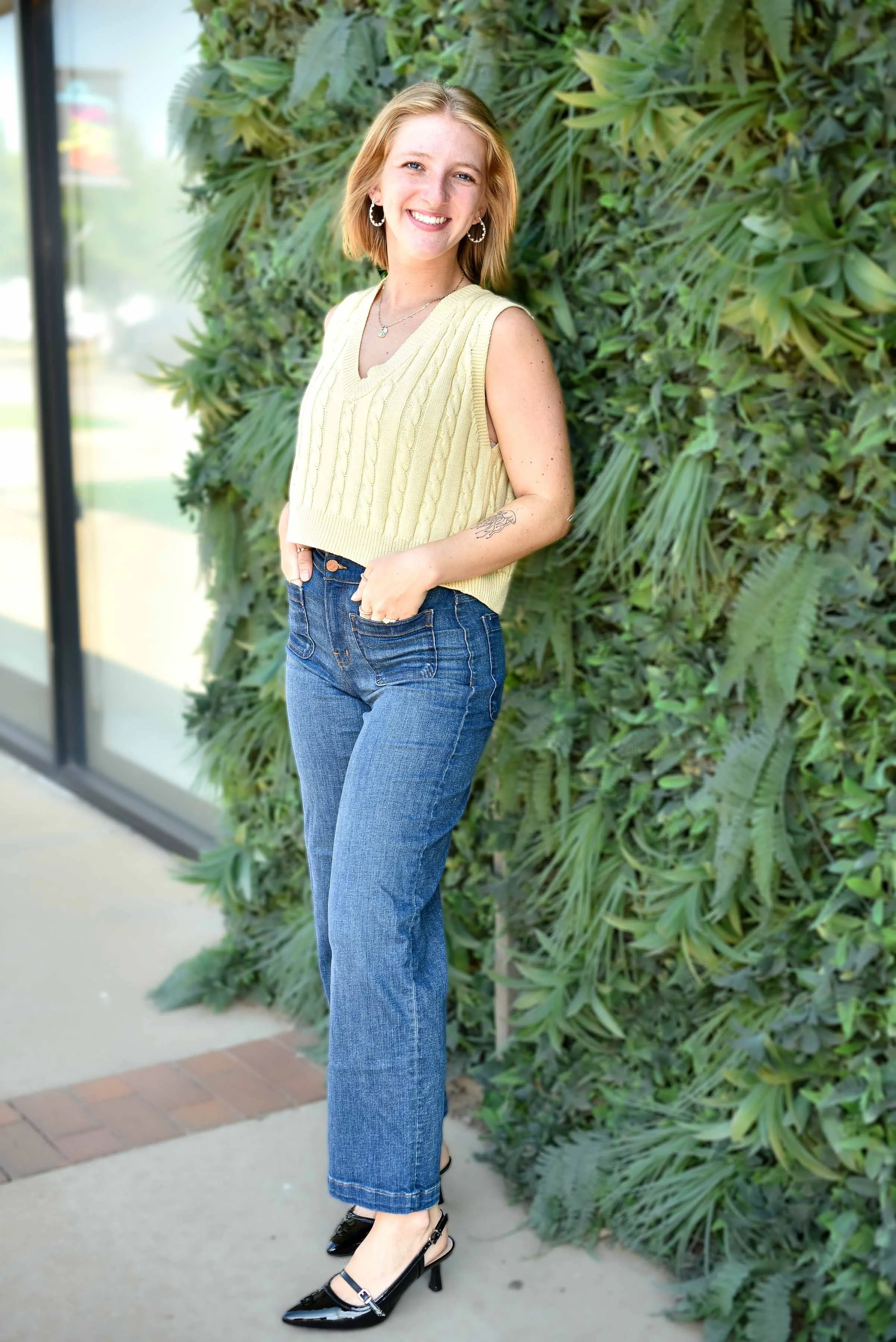 A young woman with shoulder-length blonde hair, smiling, standing outdoors near a green leafy wall. She is wearing a yellow knitted sleeveless top, blue jeans, and black high heels.