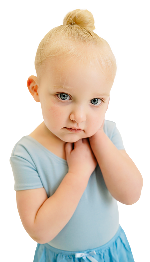 A young girl with blonde hair in a bun, wearing a light blue shirt and matching skirt, looking directly at the camera with a serious expression, her hands gently touching her neck.