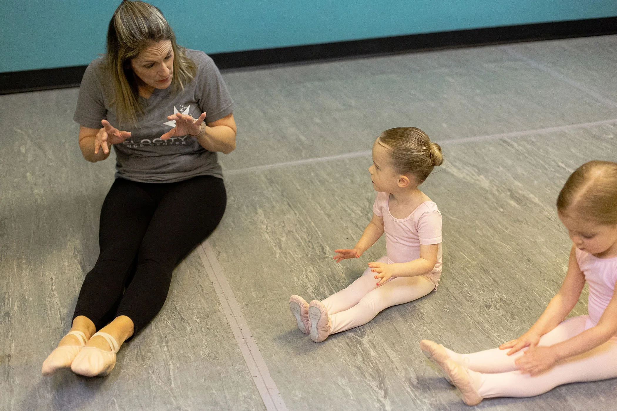 A ballet instructor sitting on the floor and talking to two young ballerinas sitting with their legs extended in front of them during a dance class.