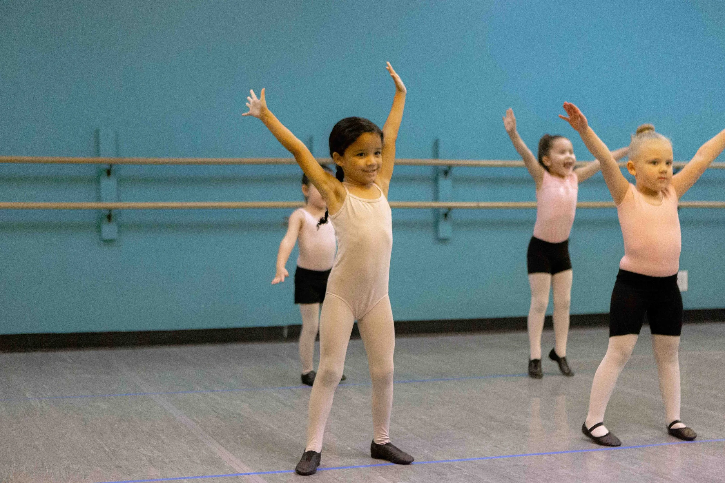 Four young girls practicing ballet in a dance studio, wearing pink leotards, black shorts, tights, and ballet shoes, with arms raised in ballet pose.