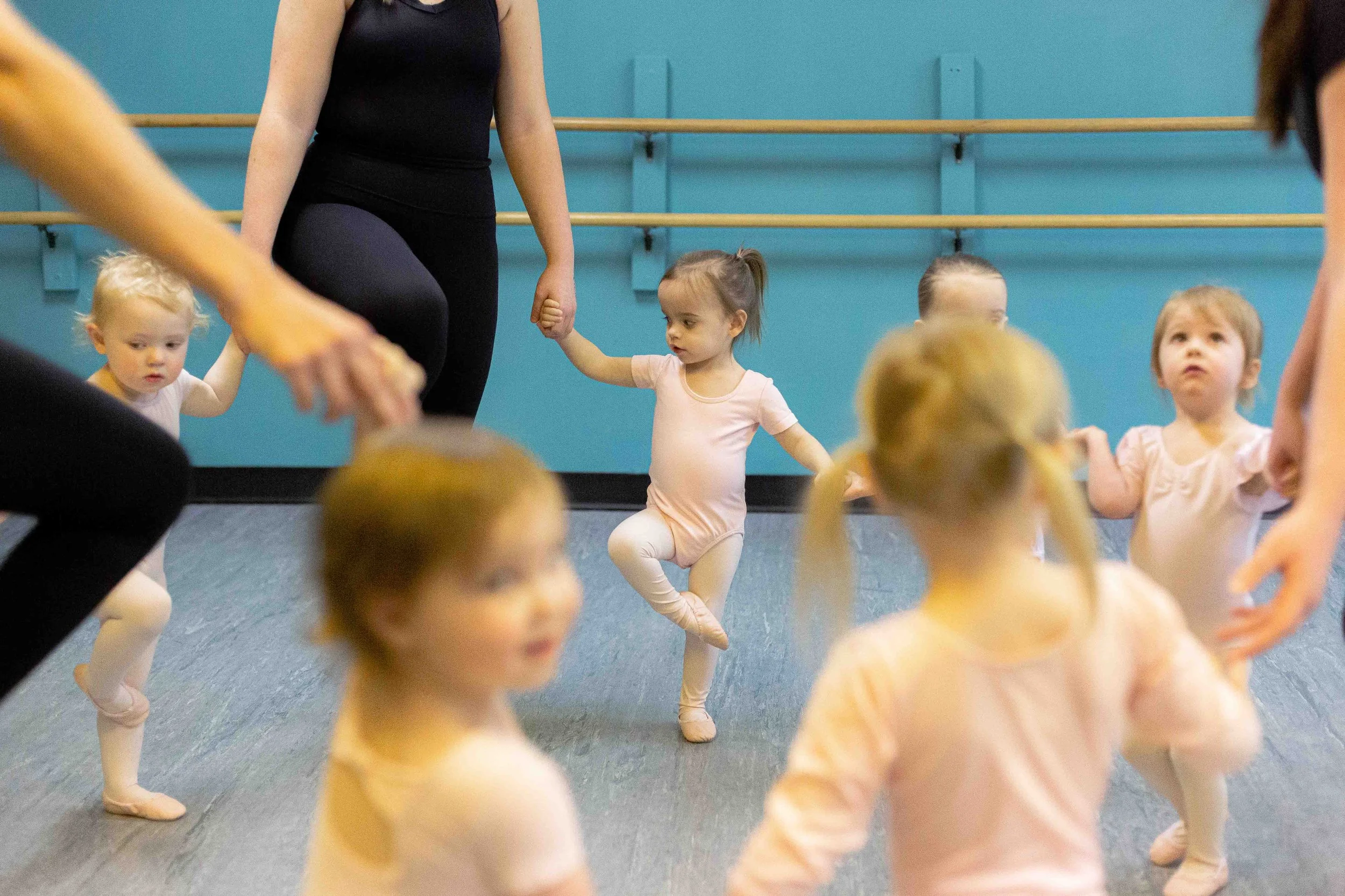 Young children participating in a ballet class, holding hands with instructors, in a dance studio with blue walls and wooden ballet barres.