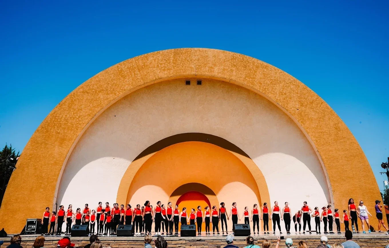 Group of performers in red tops and black pants standing on an outdoor stage with a large semi-circular yellow and white backdrop, audience watching under a clear blue sky.