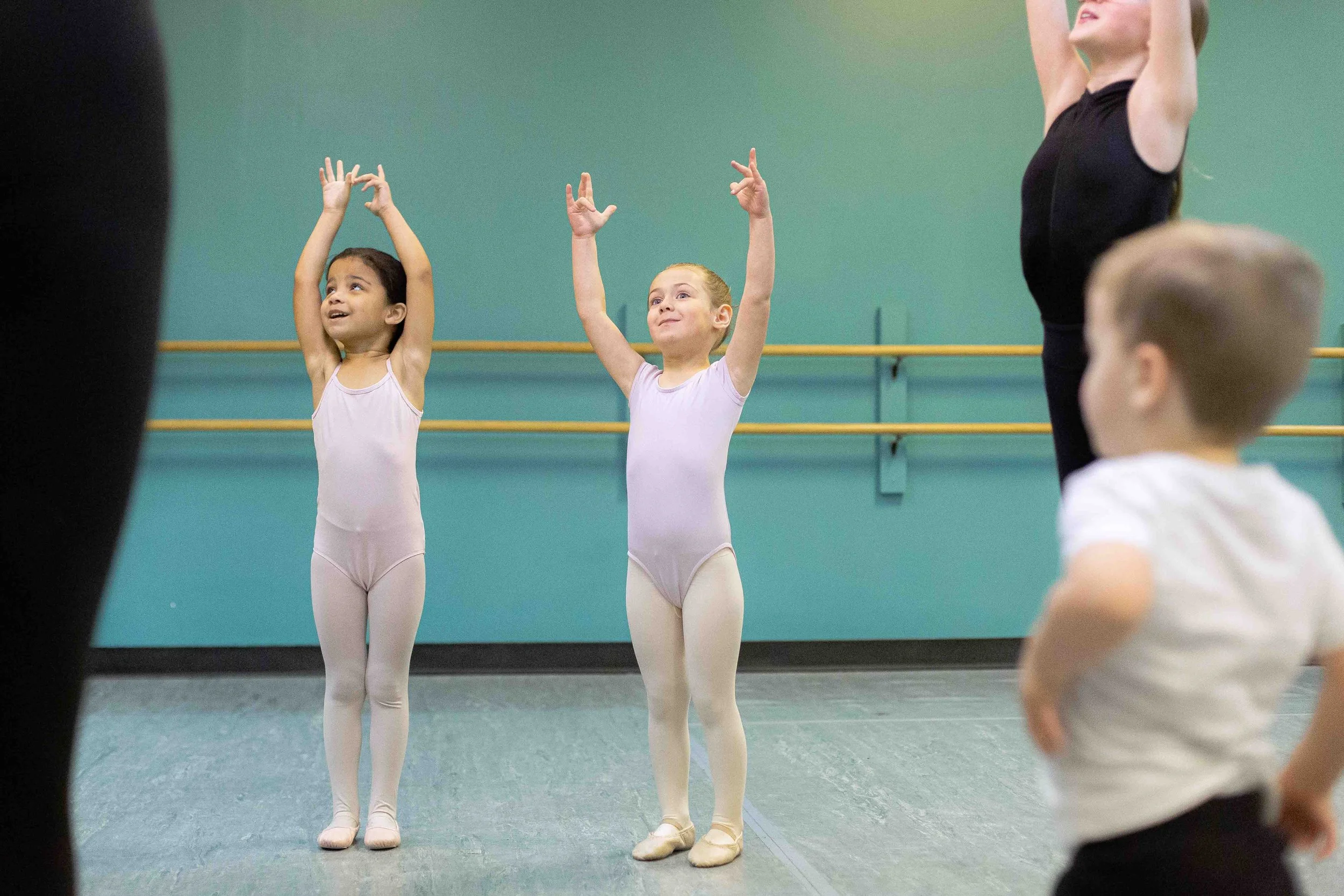 Two young girls in ballet leotards and tights practicing ballet in a dance studio with an instructor supervising, other children observing.