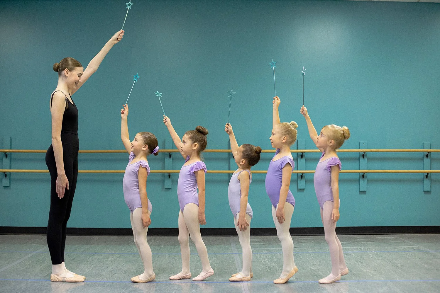 A ballet teacher with five young girls in purple leotards and white tights, holding star wands, in a dance studio with a teal wall and ballet barres.