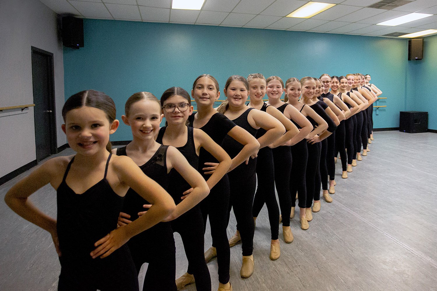 A line of young female ballet dancers in black leotards and beige ballet shoes standing in a dance studio with a teal wall and gray floor, smiling at the camera.