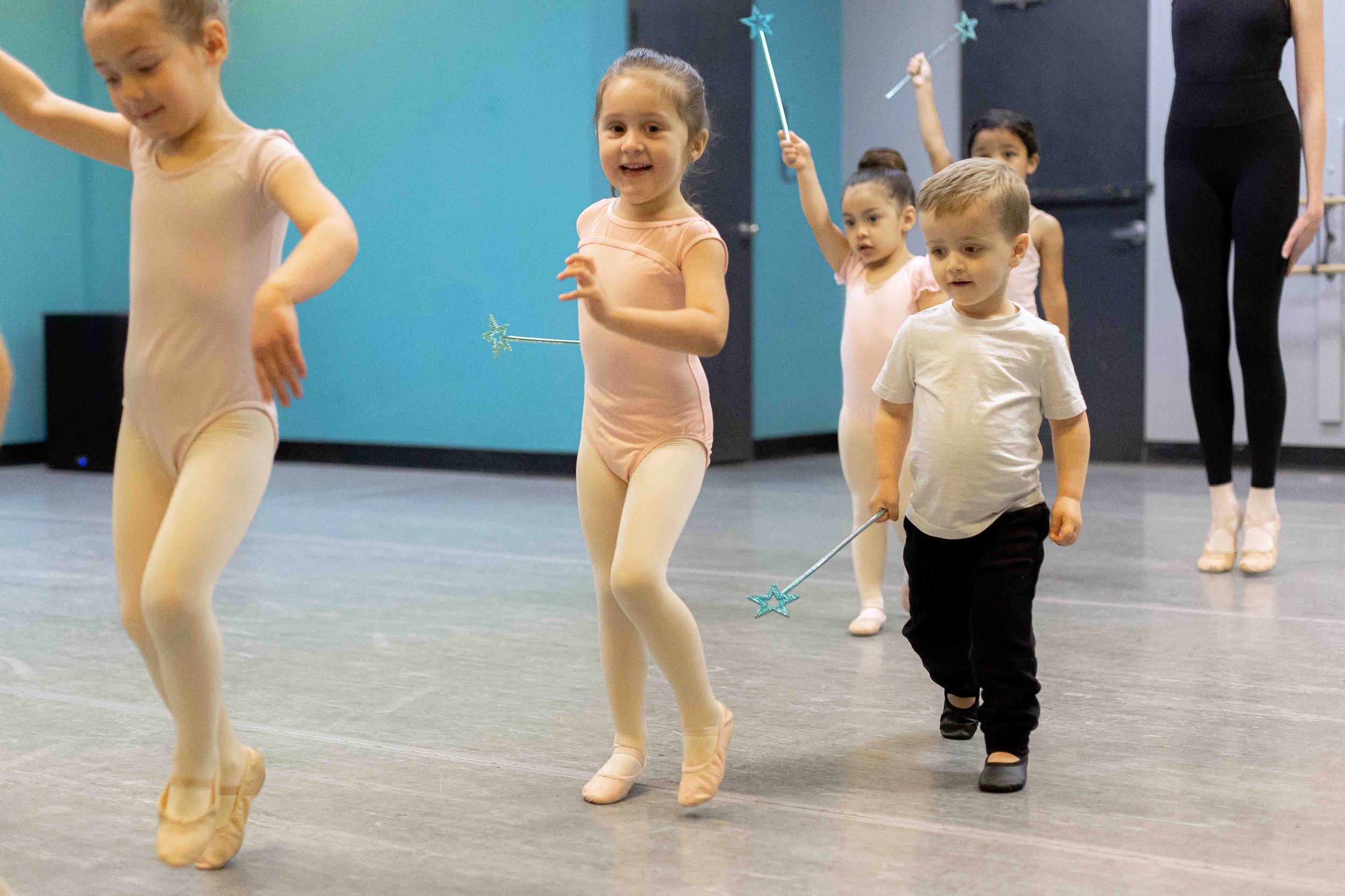 Children participating in a ballet class, holding star-shaped wands, with a ballet instructor in the background.