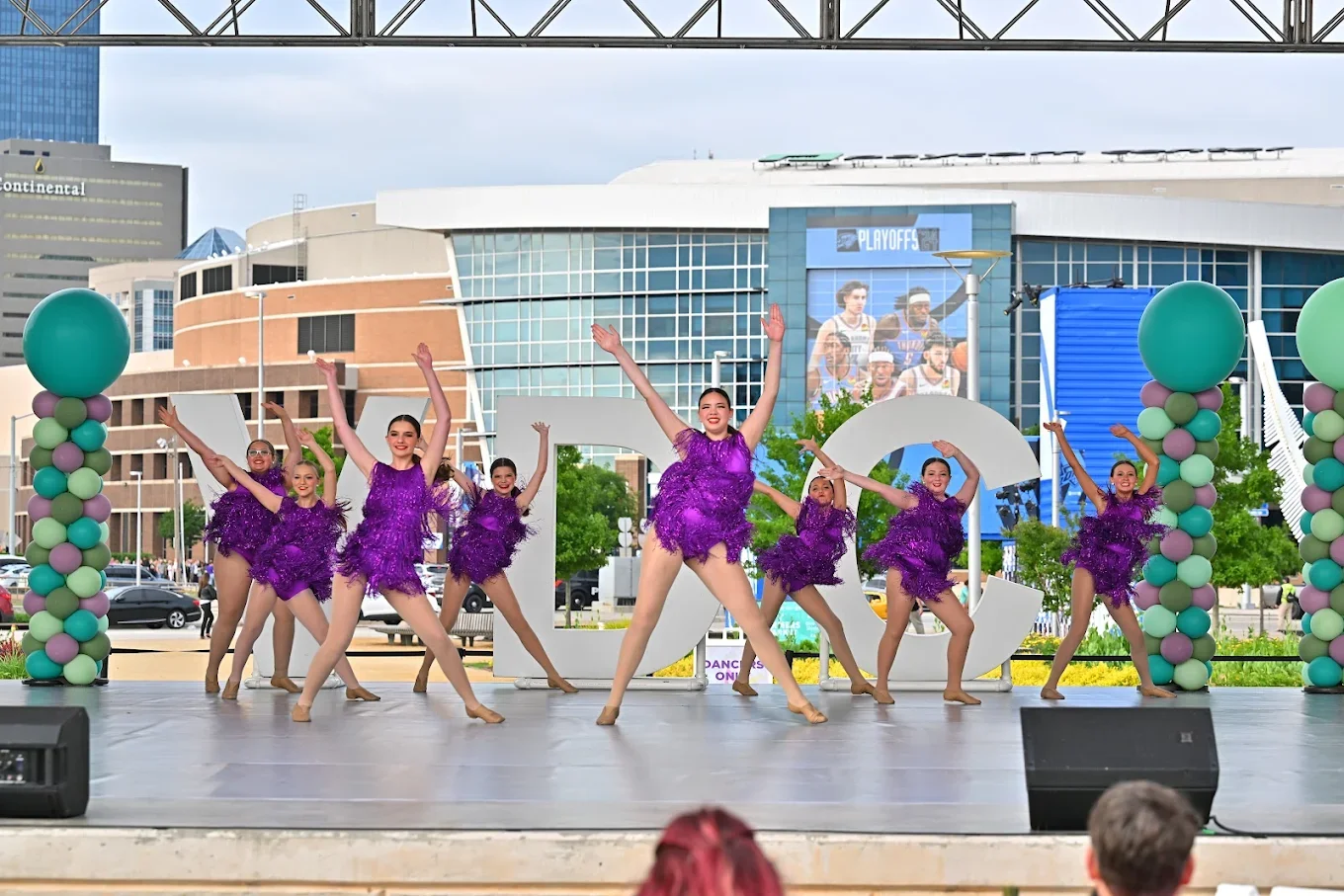 Group of young women in purple costumes performing a dance on an outdoor stage, with balloons and large decorative letters in front, in a city setting during daytime.