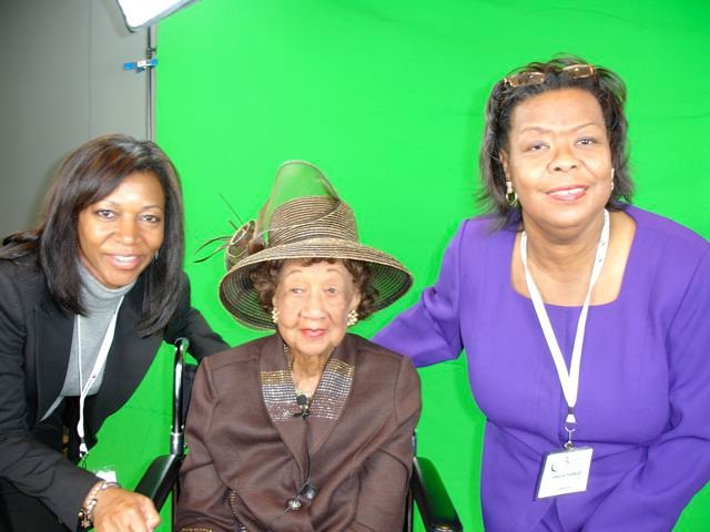 Three women posing in front of a green screen; the woman in the middle is elderly, wearing a large hat, and seated in a wheelchair. The women on either side are standing, smiling, and have on conference badges.