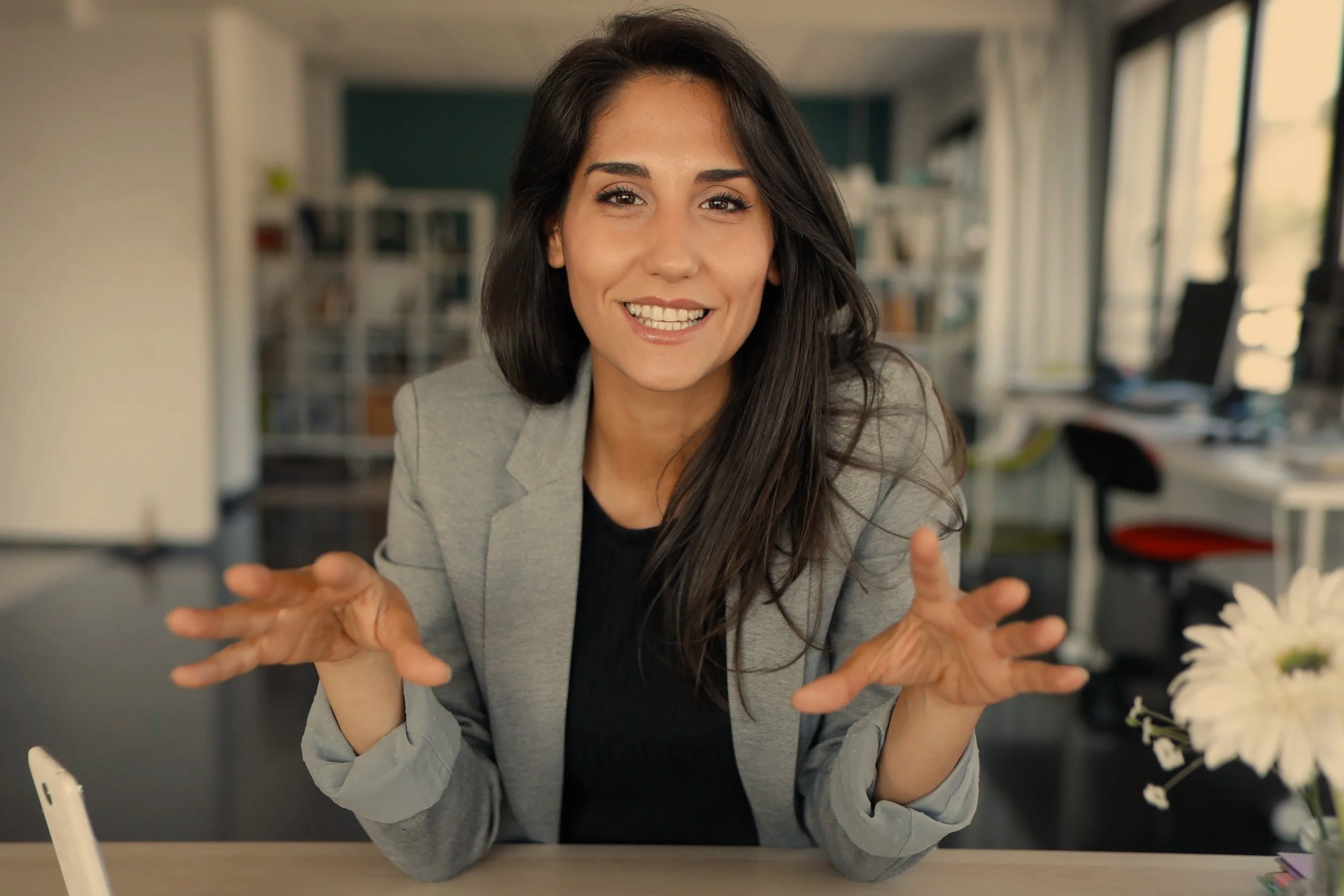 A woman with long dark hair smiling and gesturing with her hands while sitting at a desk in an office.