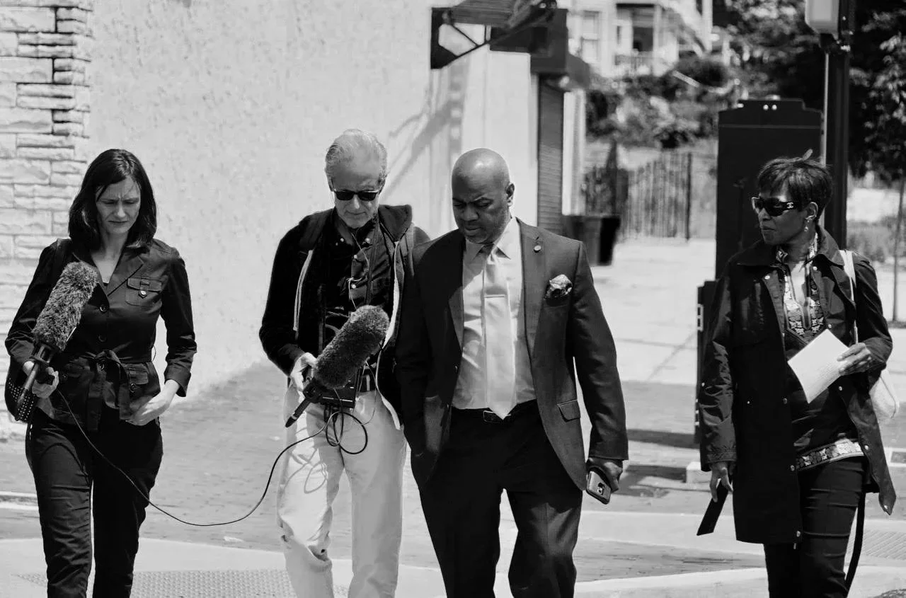 Group of four people walking outdoors with a wall and trees in the background. Two women and two men, some carrying papers and a microphone, appear to be involved in a news or interview setting.