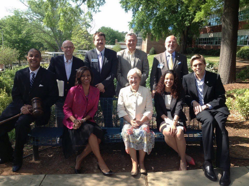 Group of ten professionally dressed people, sitting and standing on and behind a blue bench outdoors on a sunny day, with trees and a building in the background.